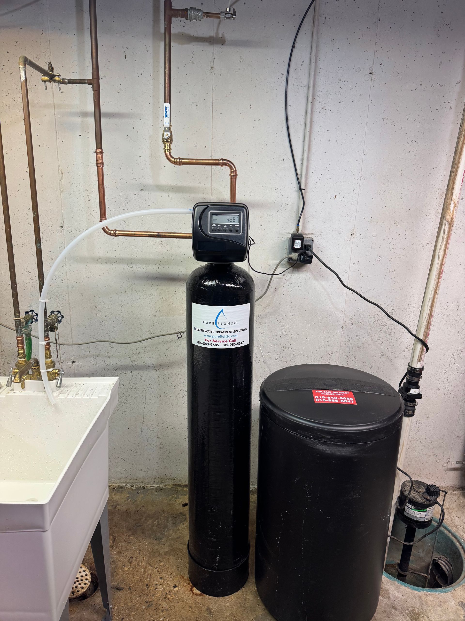 Water softener system in a basement tall black tank, salt storage, copper pipes, and a utility sink.