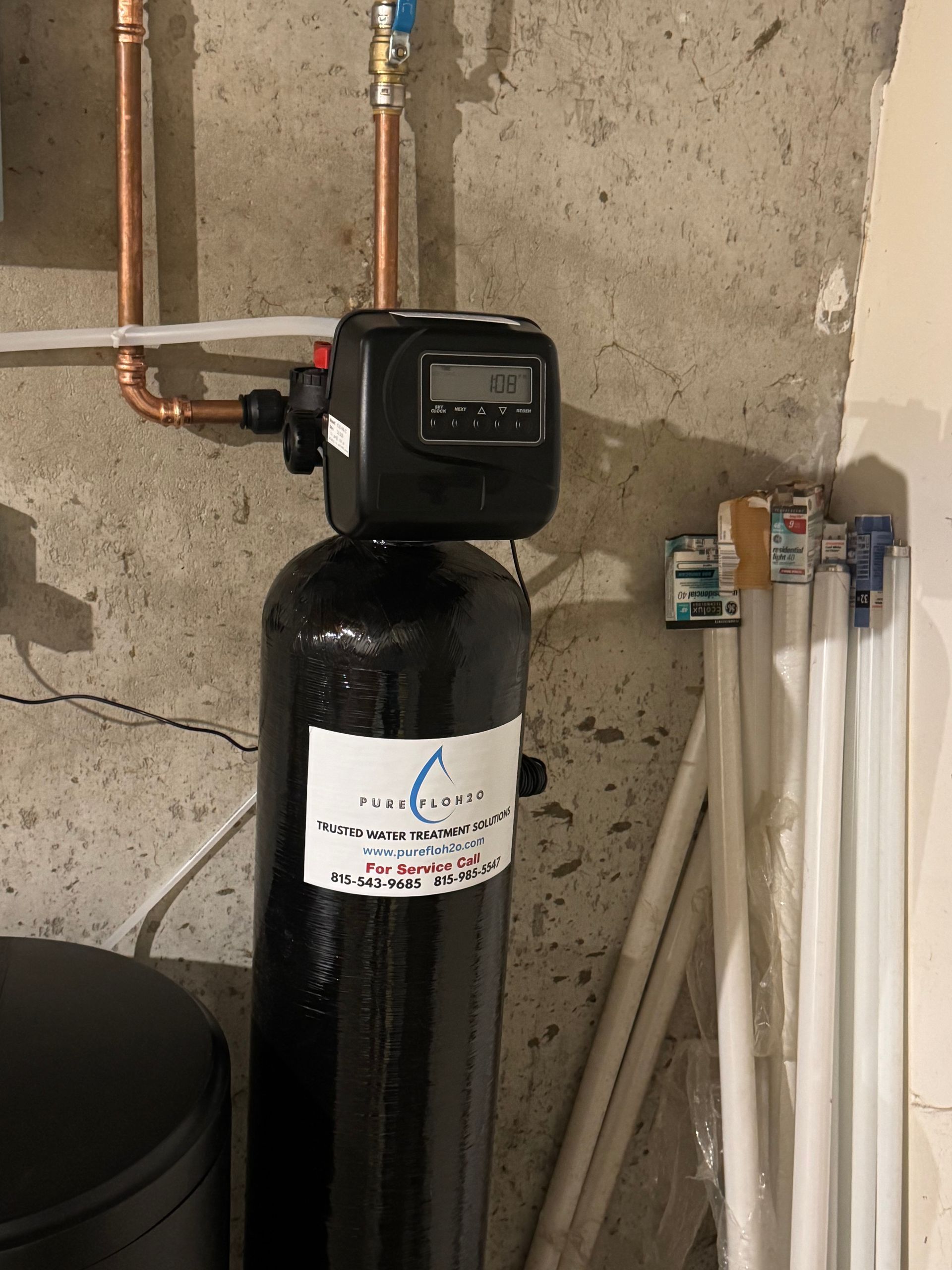 Black water softener unit with digital display and copper pipes against a concrete wall.