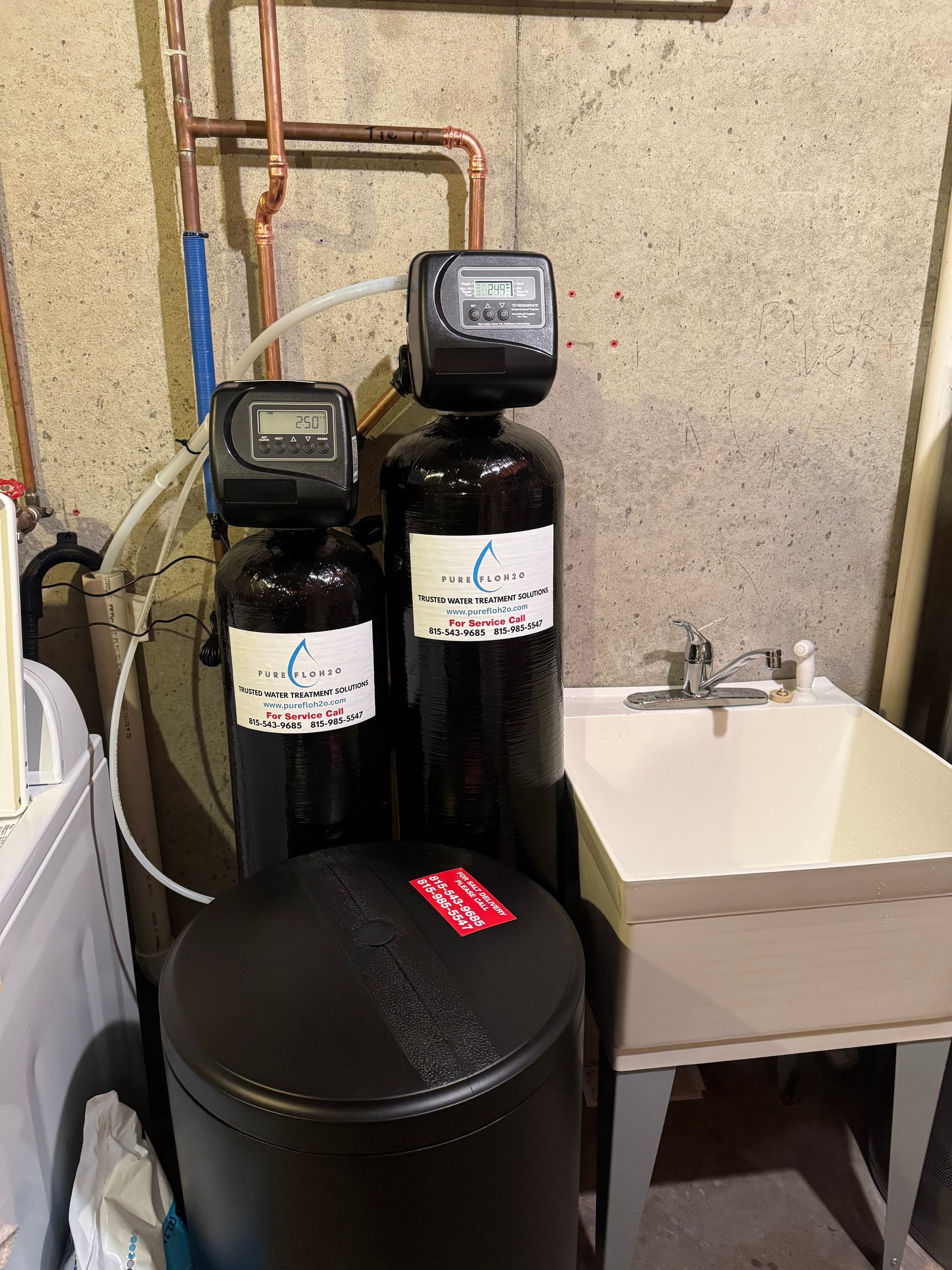 Water softener system with two black tanks, a salt tank, and copper pipes, next to a utility sink.