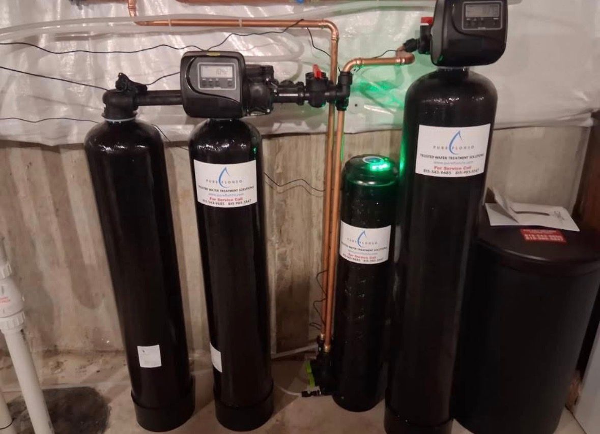 Four black water filtration tanks with digital control heads and copper piping against a basement wall. 