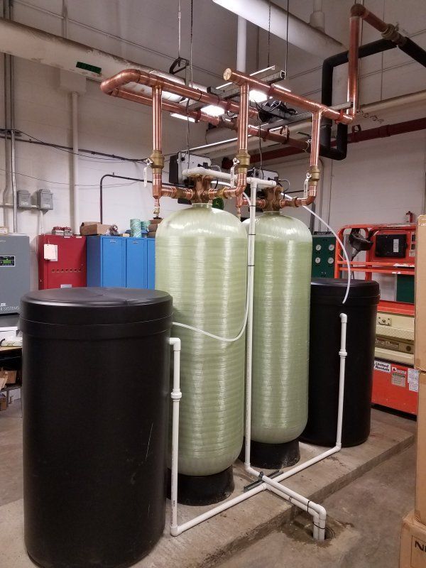 Two large, light green water treatment tanks connected by copper pipes, flanked by two black brine tanks in a facility. 