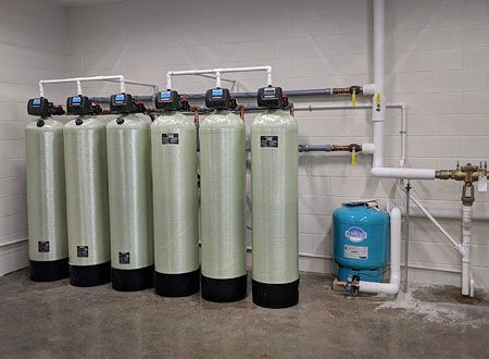 A row of six tall, light green industrial water treatment tanks with black bases and plumbing against a white wall. 