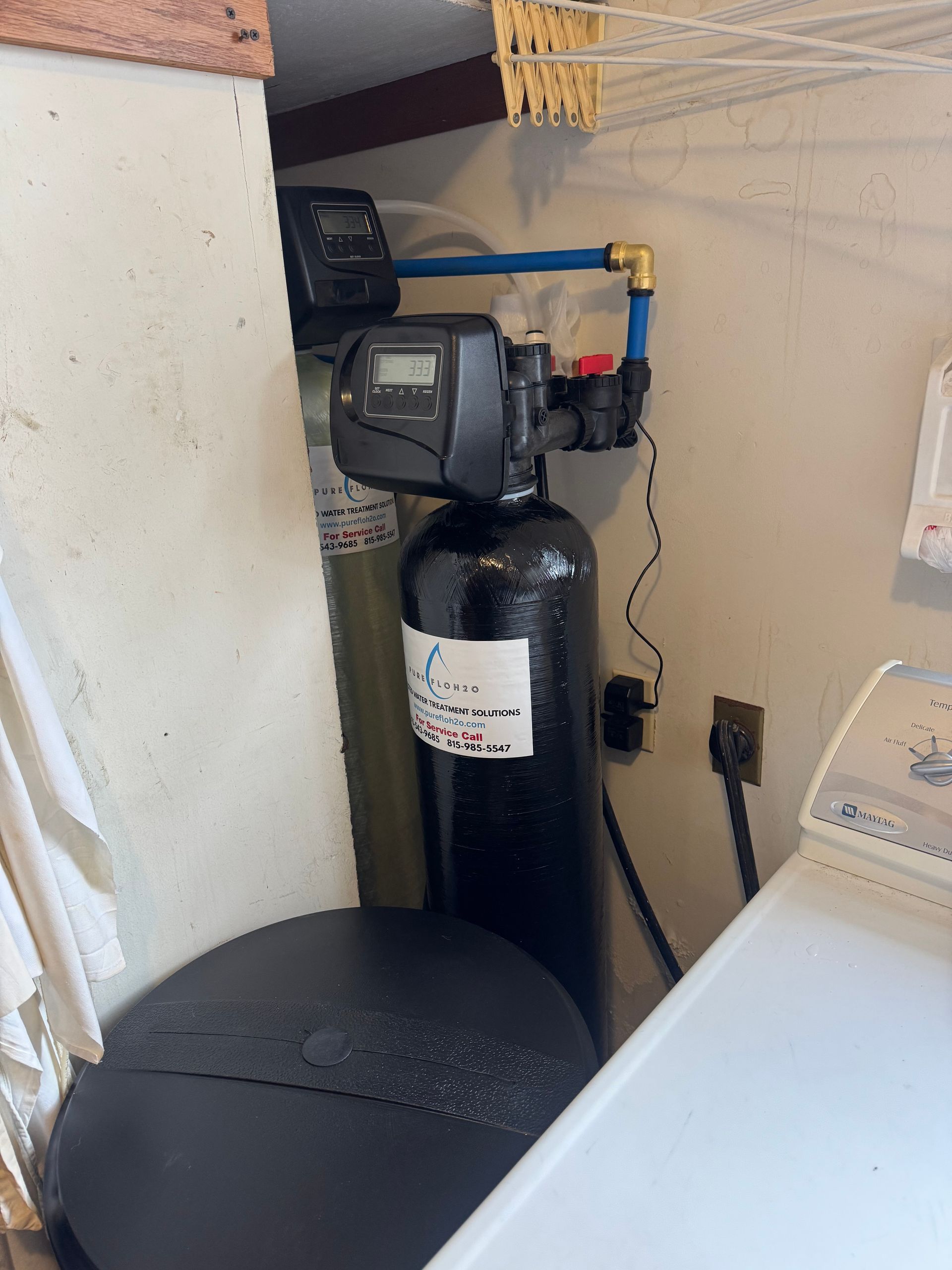Two water treatment system tanks with digital controls installed in a small closet next to a washing machine. 