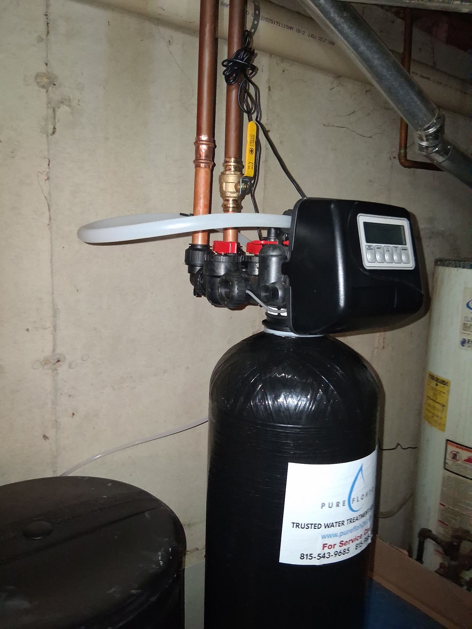 A water softener system connected to copper pipes, featuring a black cylindrical tank and a digital control panel. 