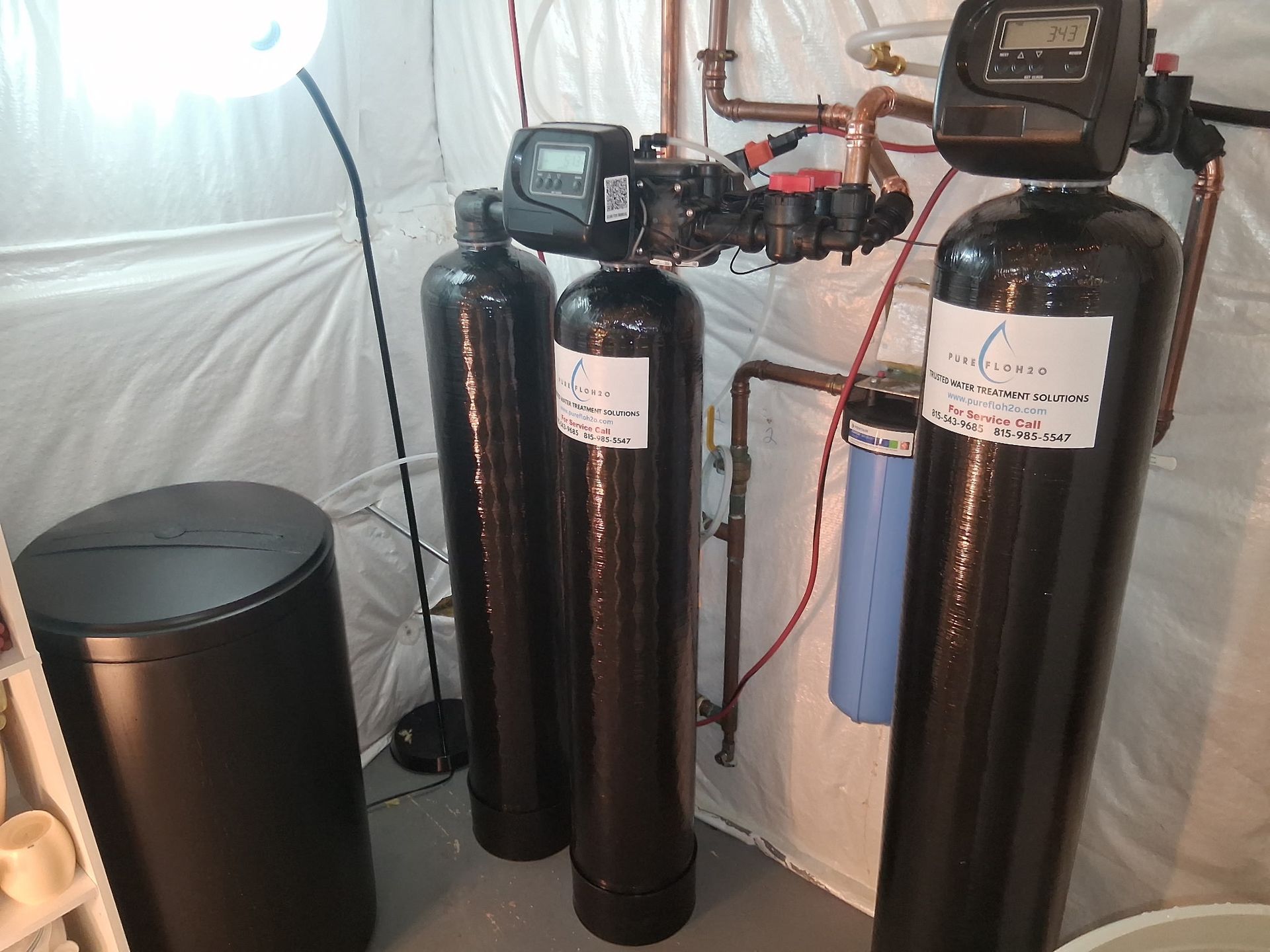 Water filtration system in a basement.  Multiple black tanks and a blue filter connected by copper pipes.