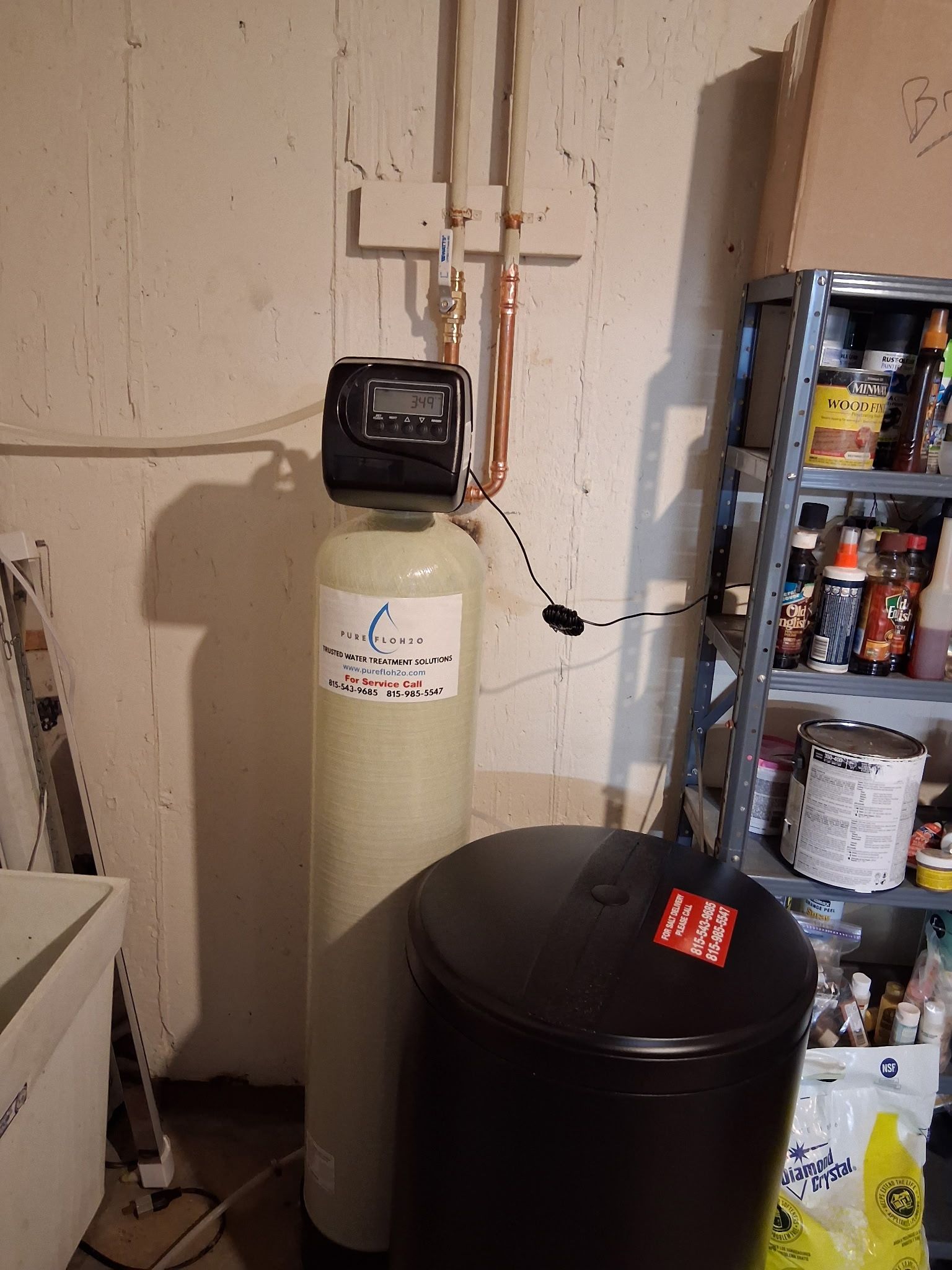 A beige water softener tank with a digital control head next to a black brine tank in a basement. 
