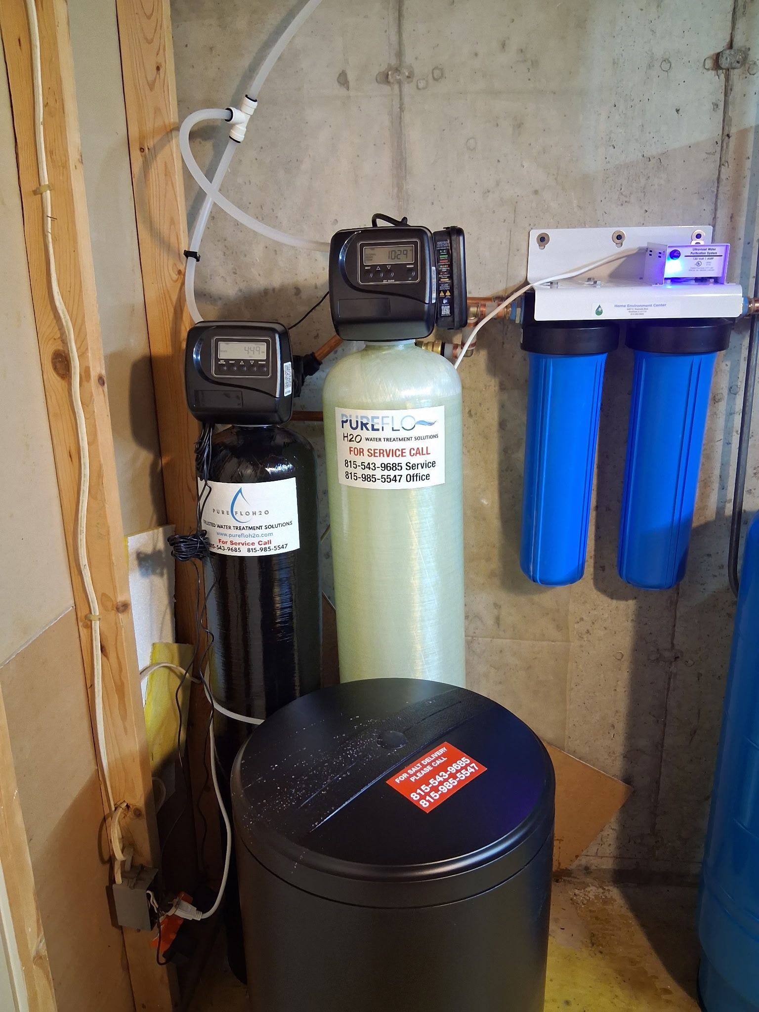 A water filtration system in a basement featuring two tanks, a brine barrel, and two wall-mounted blue canister filters. 