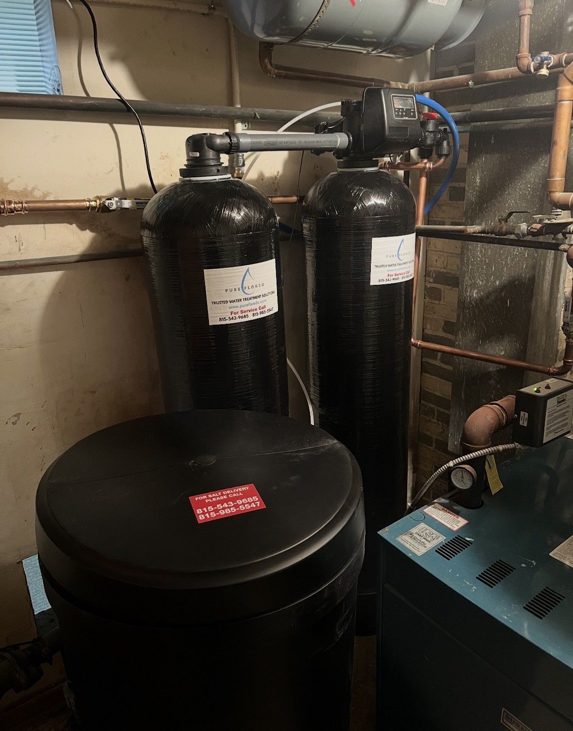 Two black water softener tanks in a basement utility room with pipes and a blue appliance
