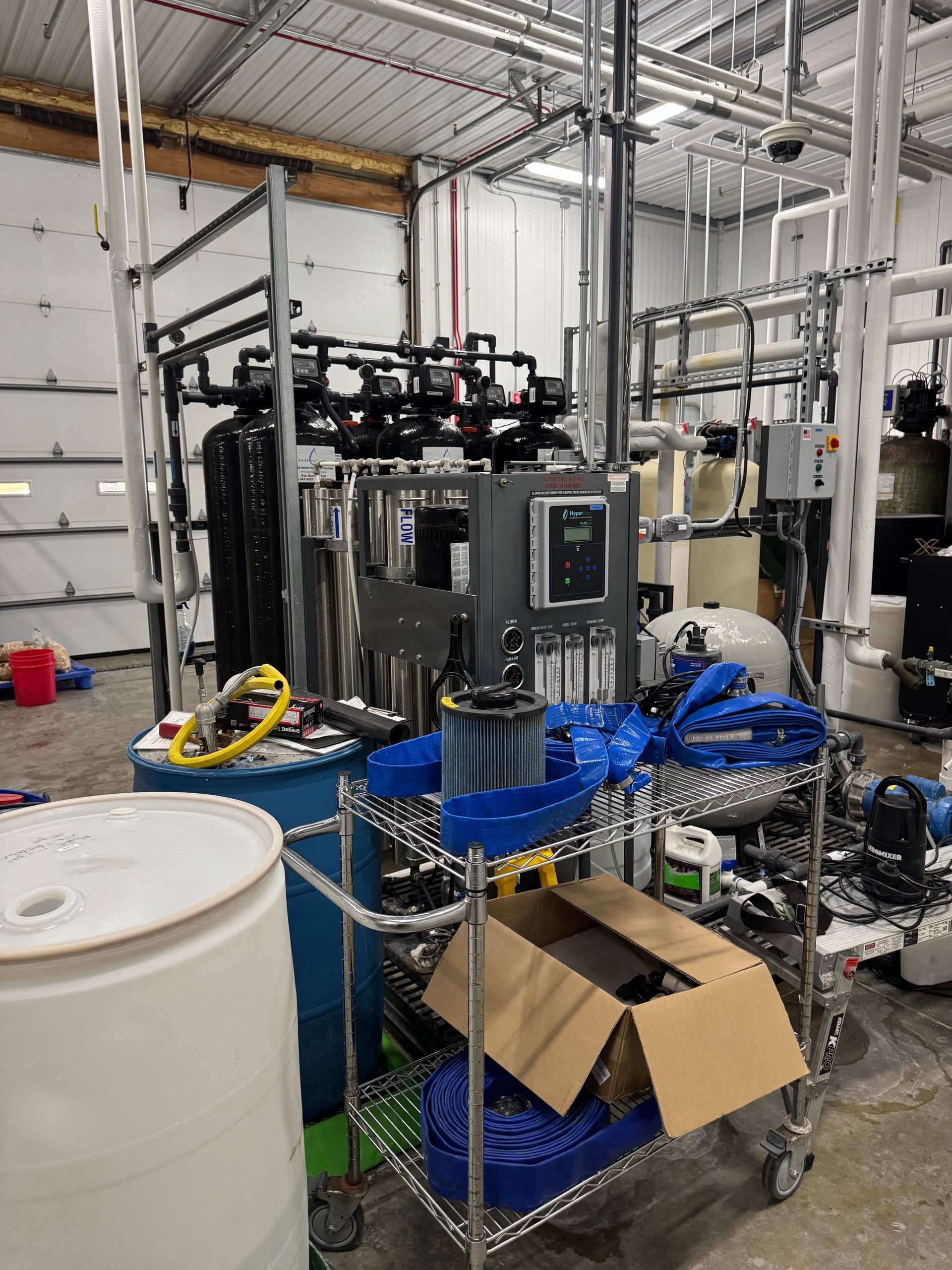 Industrial machinery in a factory, with blue hoses, a control panel, and a wheeled cart with a box.