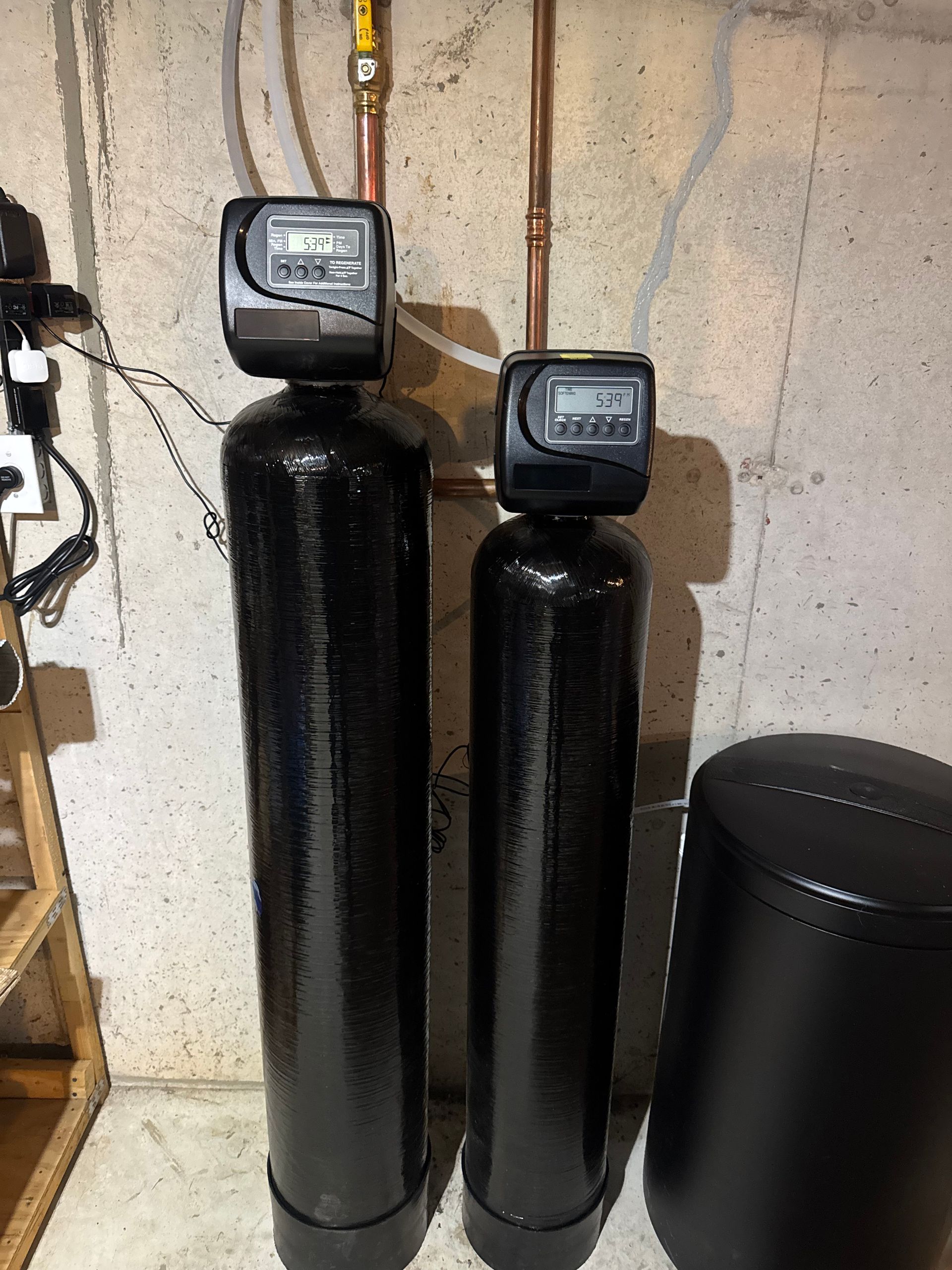 Two black water treatment tanks with digital displays, connected to copper pipes, and a salt tank against a concrete wall.