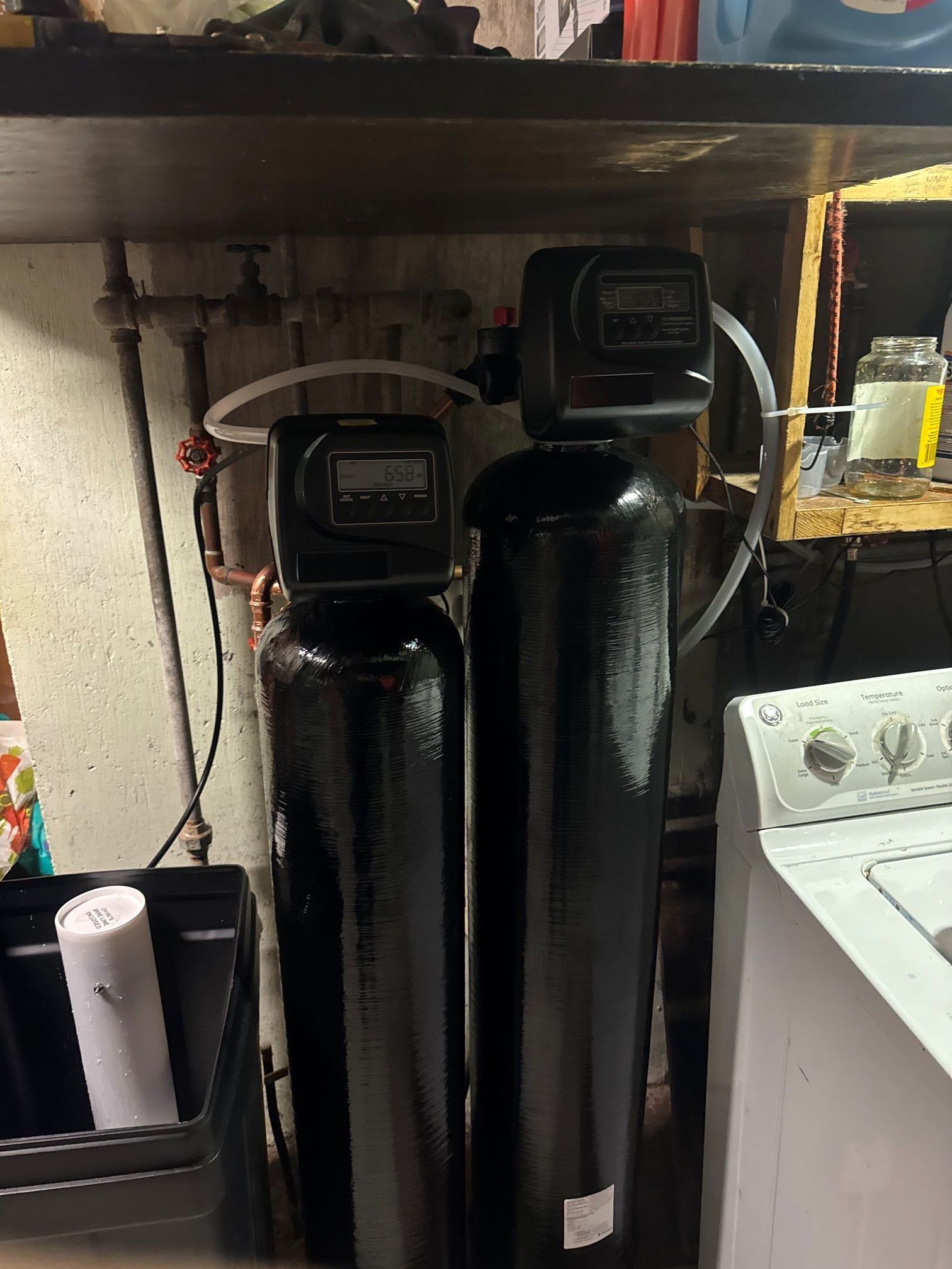 Two black water filtration tanks with digital displays, connected by tubing, beside a washing machine.