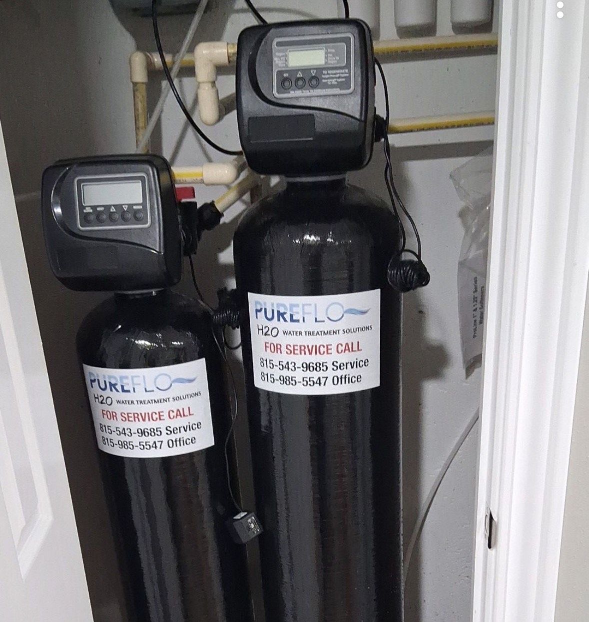 Two black water filtration systems with digital displays, plumbing, and a PureFlow label inside a utility closet.