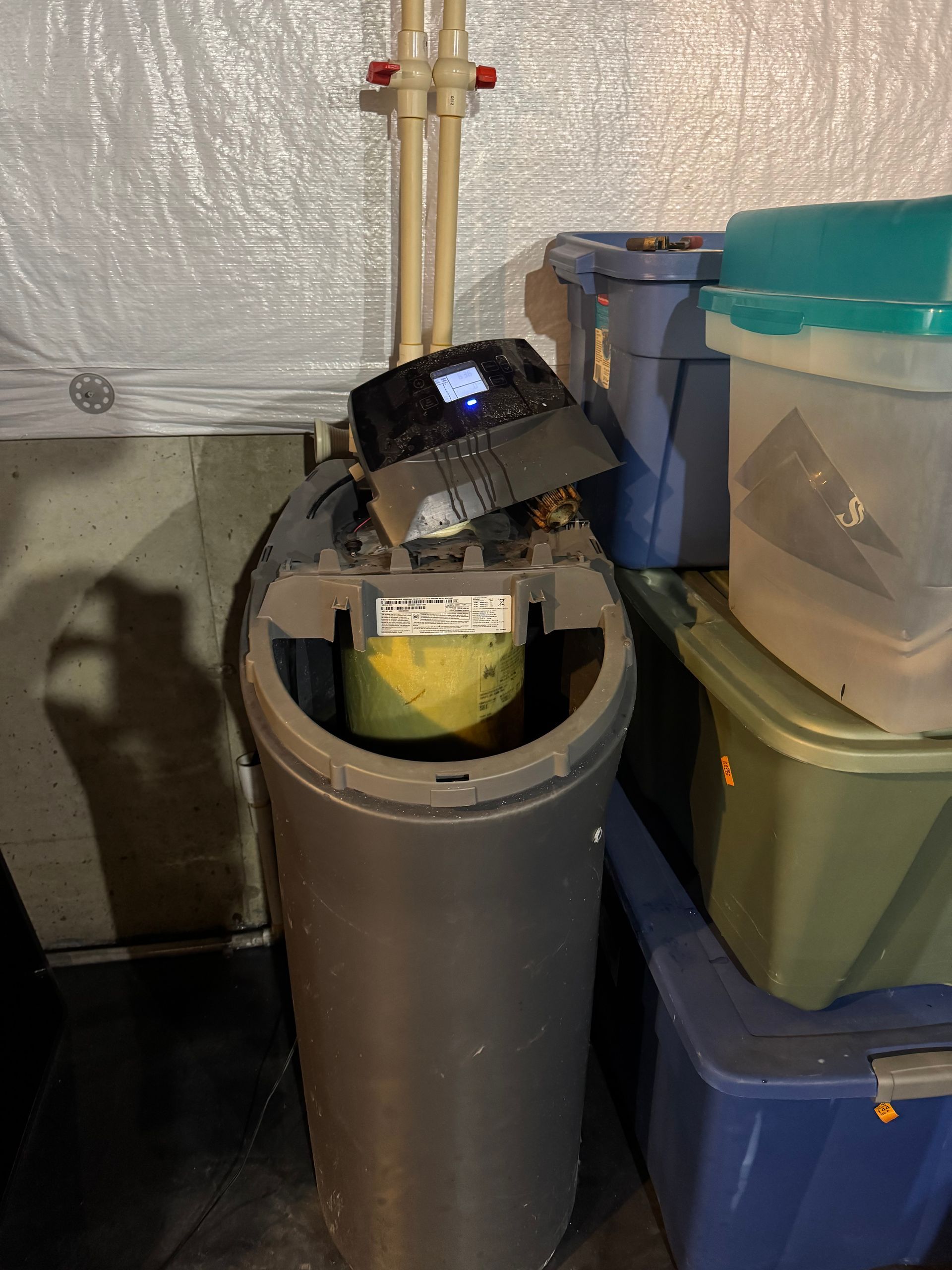 Water softener tank with control panel, pipes, and storage bins in a basement.
