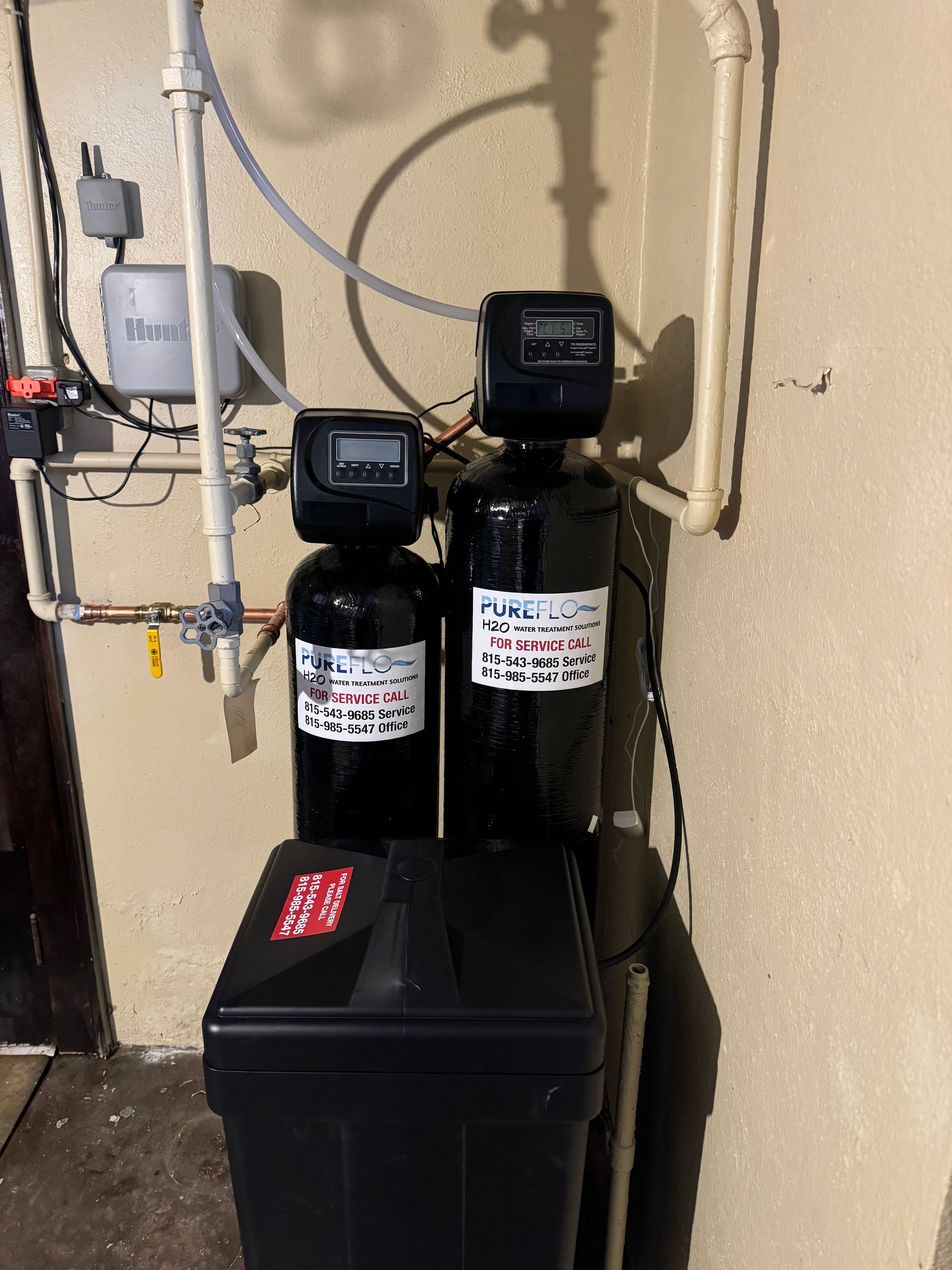 Water filtration system with two black tanks and a salt storage tank against a beige wall.