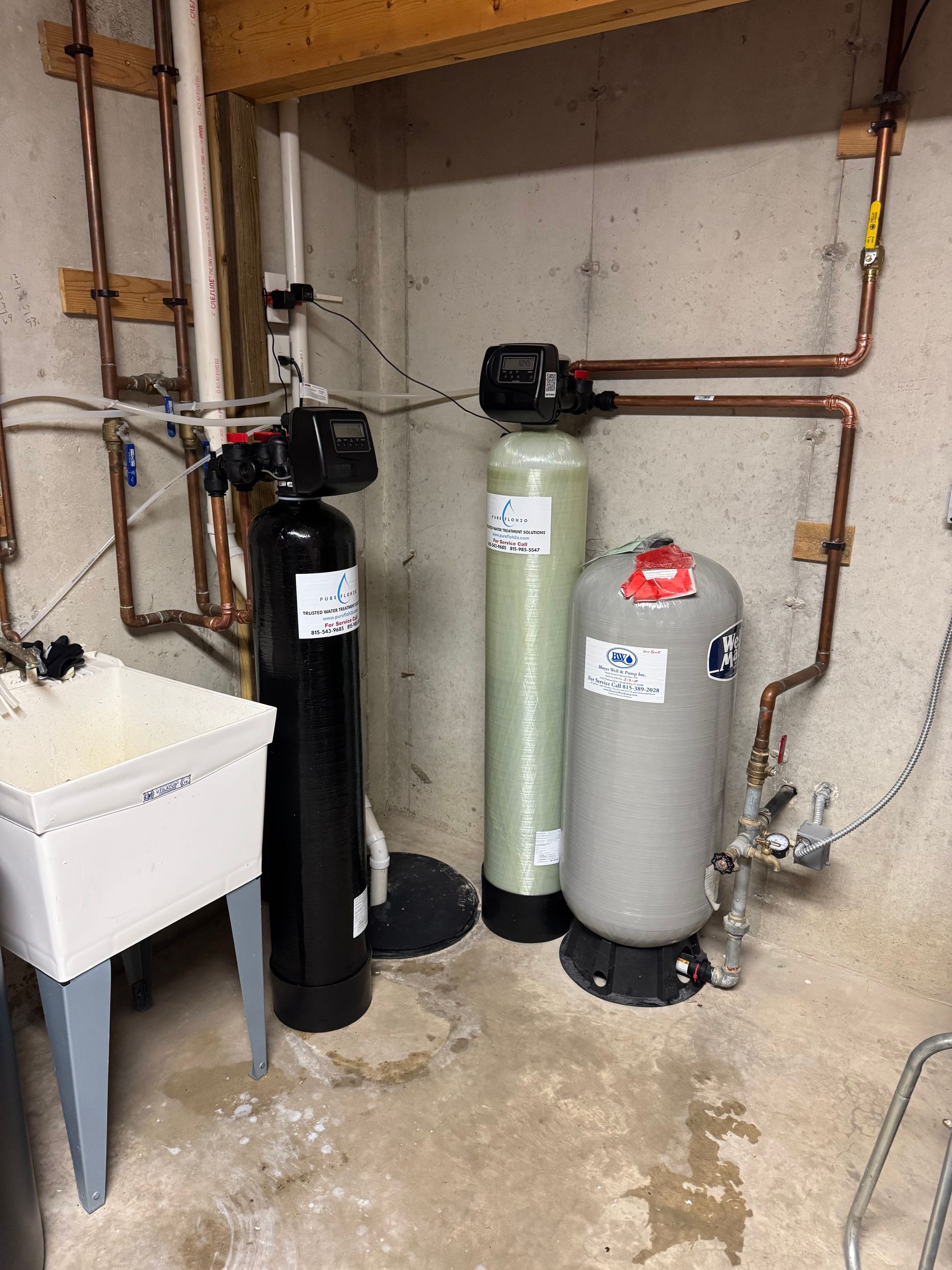 Plumbing system in a basement with water softener, filter, and pressure tank; copper pipes, concrete walls, and a utility sink.