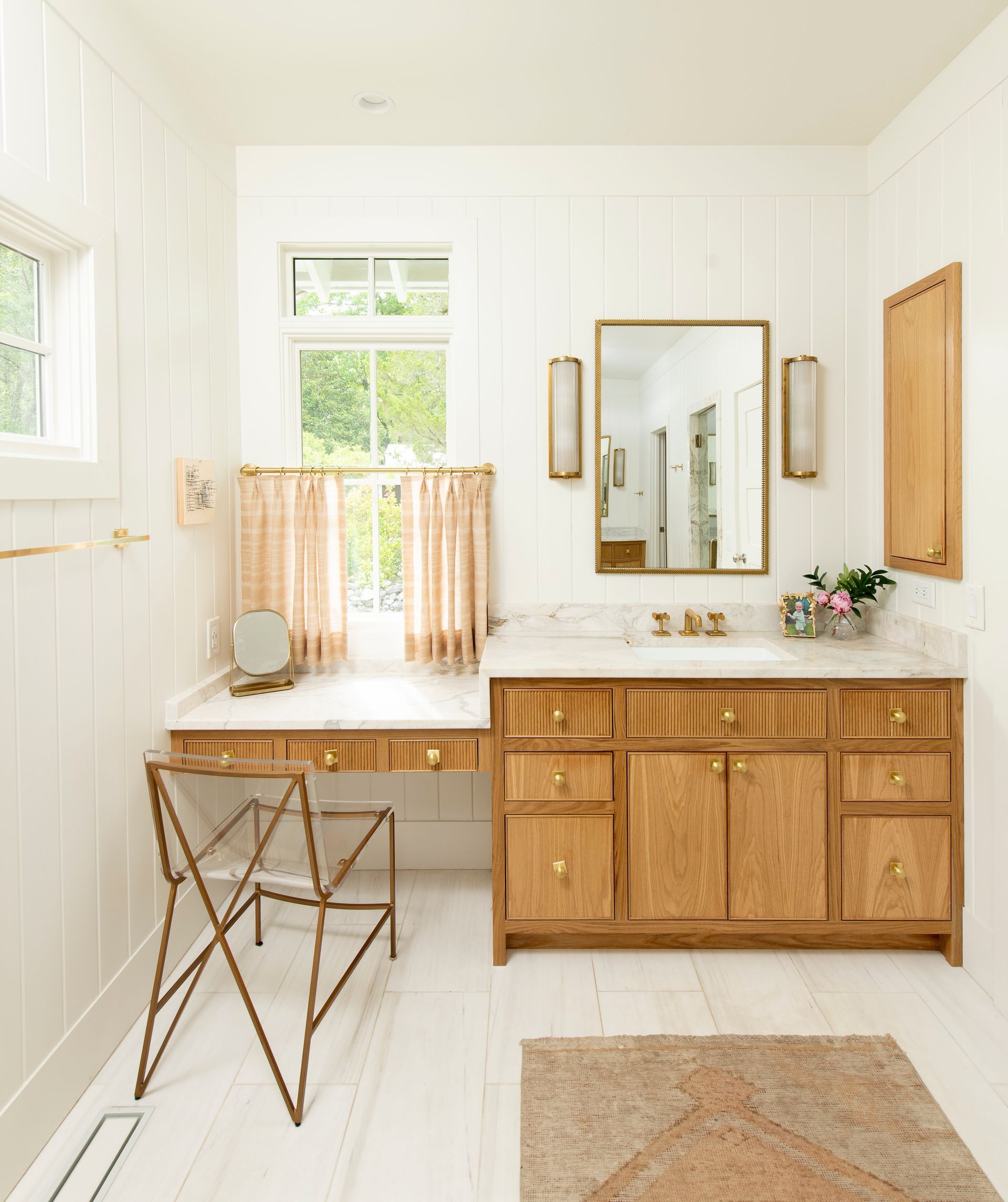 A bright bathroom with a wooden vanity, gold accents, and a small vanity desk, with a window letting in natural light.