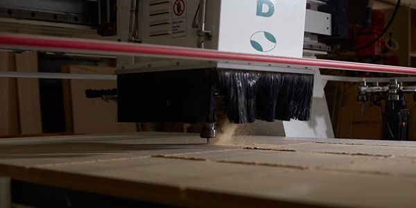A CNC router cutting wood. The machine is white with a black dust collection shroud. Sawdust is visible.