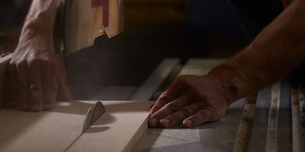 A person using a table saw to cut a piece of wood. The person's hands are visible, guiding the wood towards the blade.
