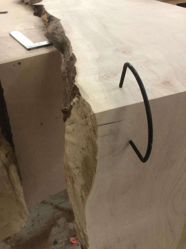 A close-up of a light-colored wooden slab with a natural edge. A bent, black metal rod is inserted for support.