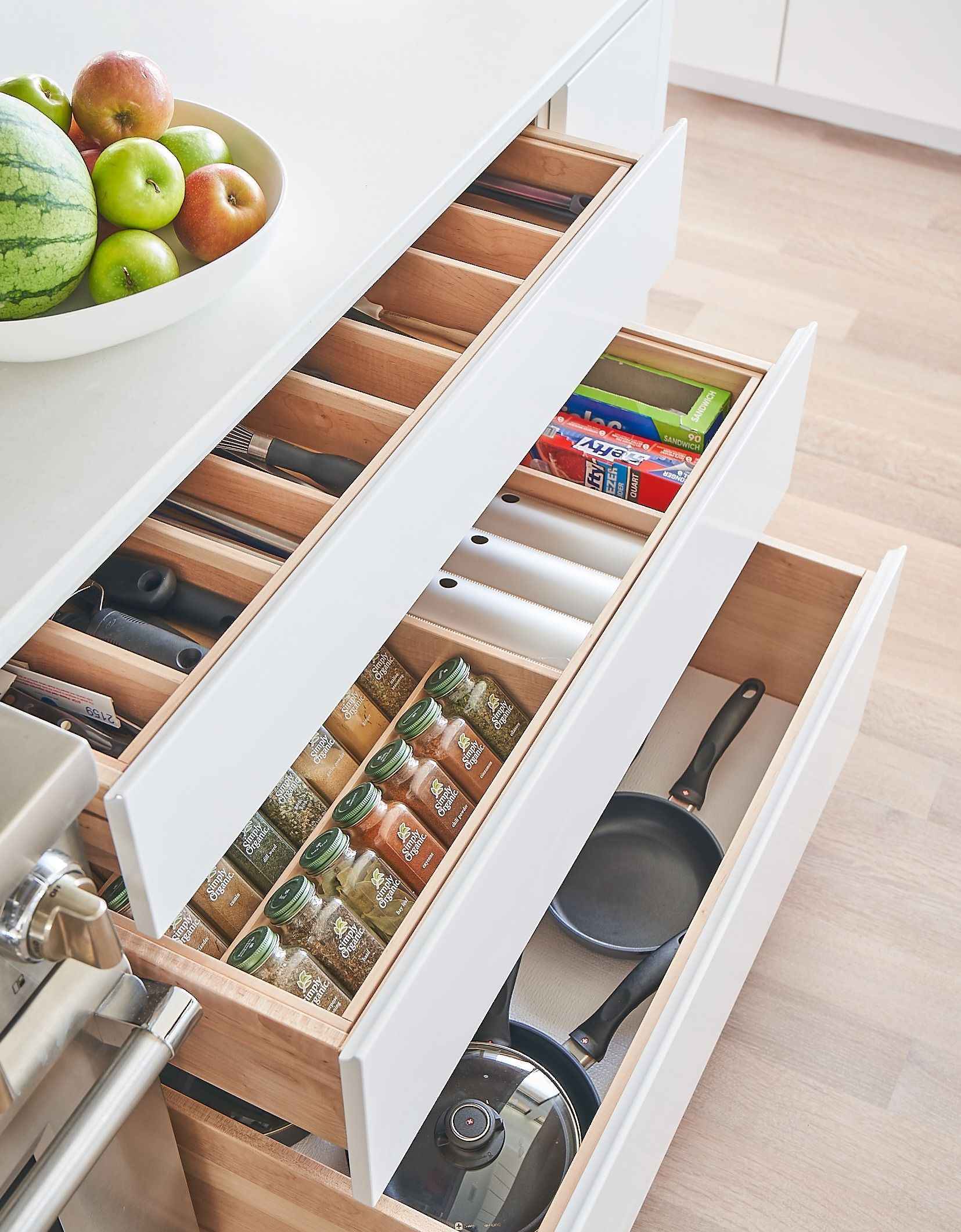 Open kitchen drawers with organized utensils, spices, and cookware. A fruit bowl is to the side.