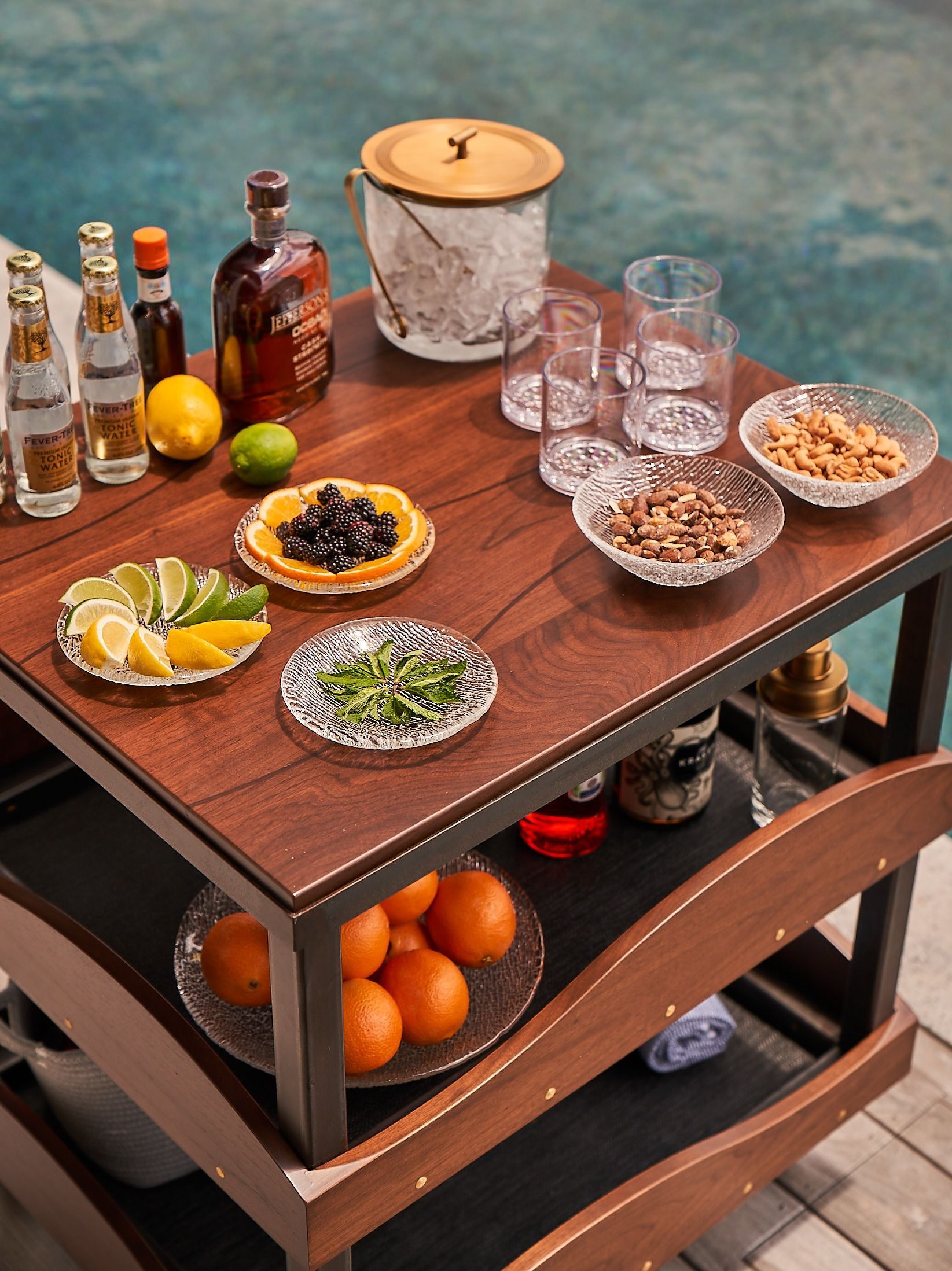 A wooden bar cart by a pool, stocked with drinks, garnishes, and snacks, including liquor bottles, citrus fruits, and nuts.