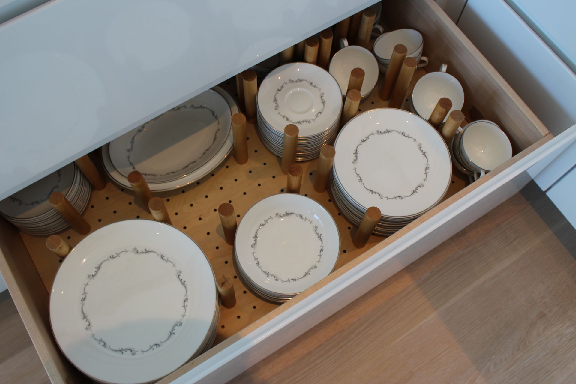 A drawer filled with stacks of white dishes separated by wooden pegs. The drawer is inside a light wood cabinet.