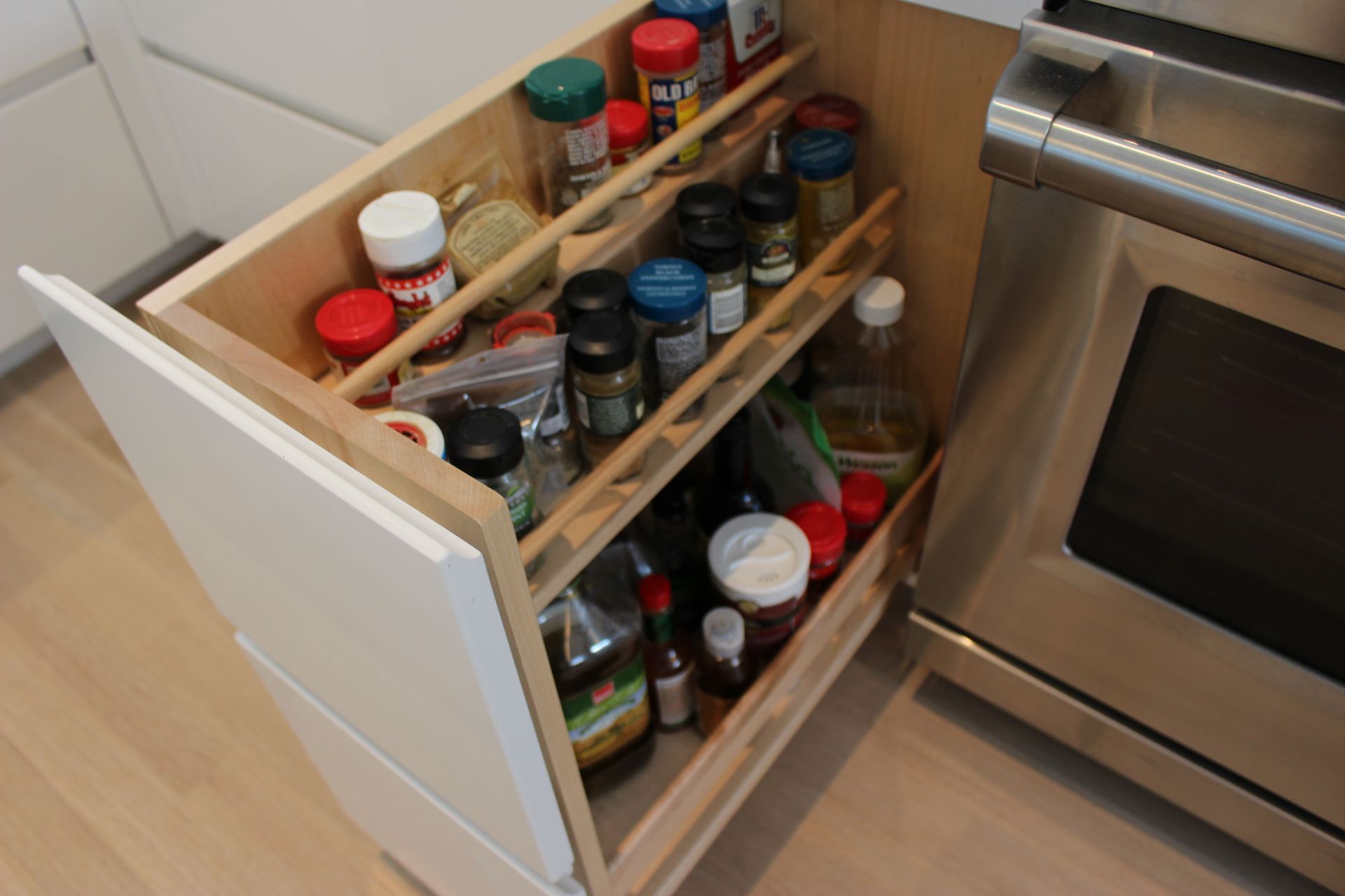 A pull-out spice rack in a white kitchen cabinet next to a stainless steel oven. The rack is filled with various spice jars.