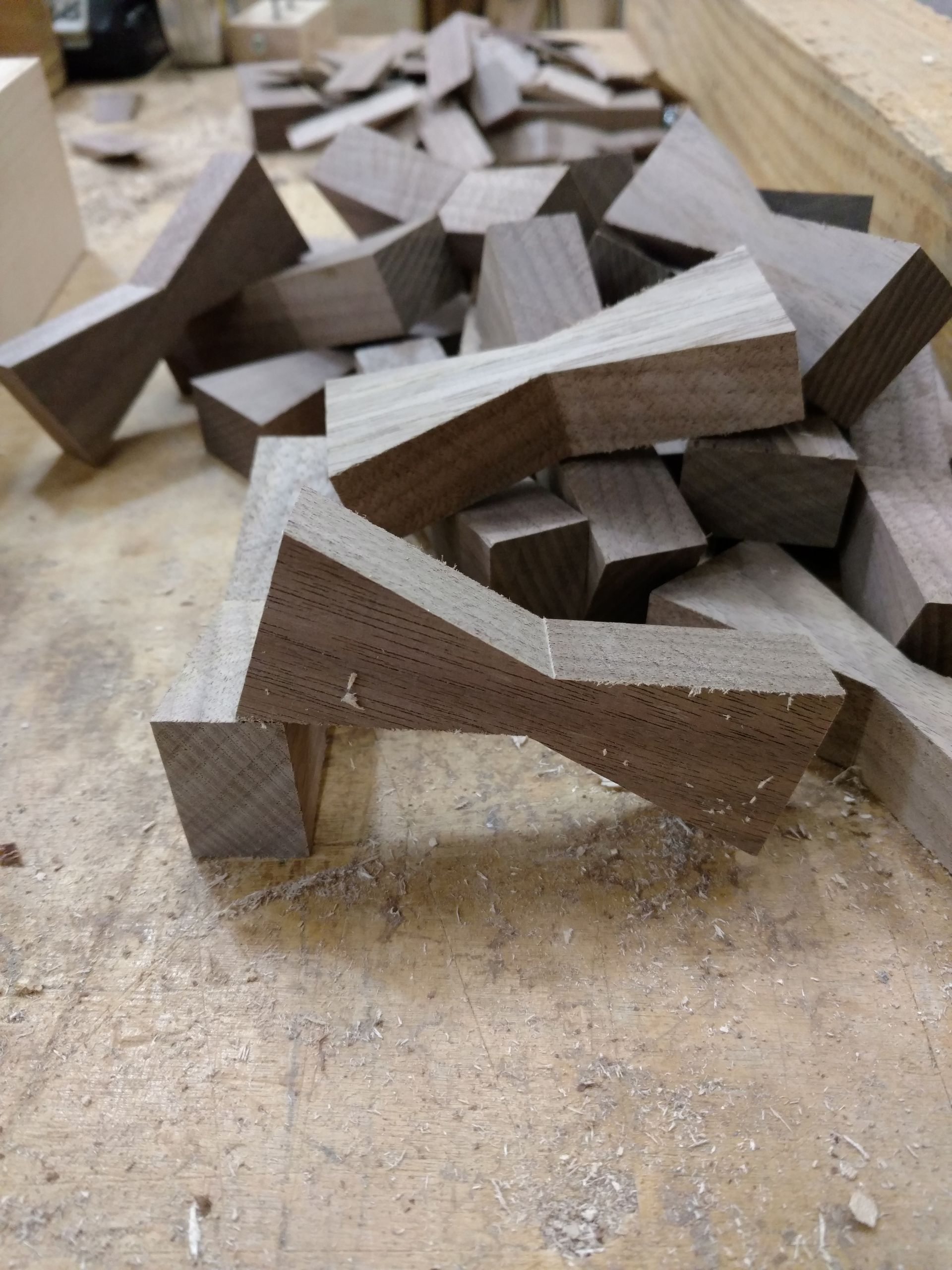 Pile of dark-brown wooden bowtie-shaped pieces scattered on a dusty wooden workbench.