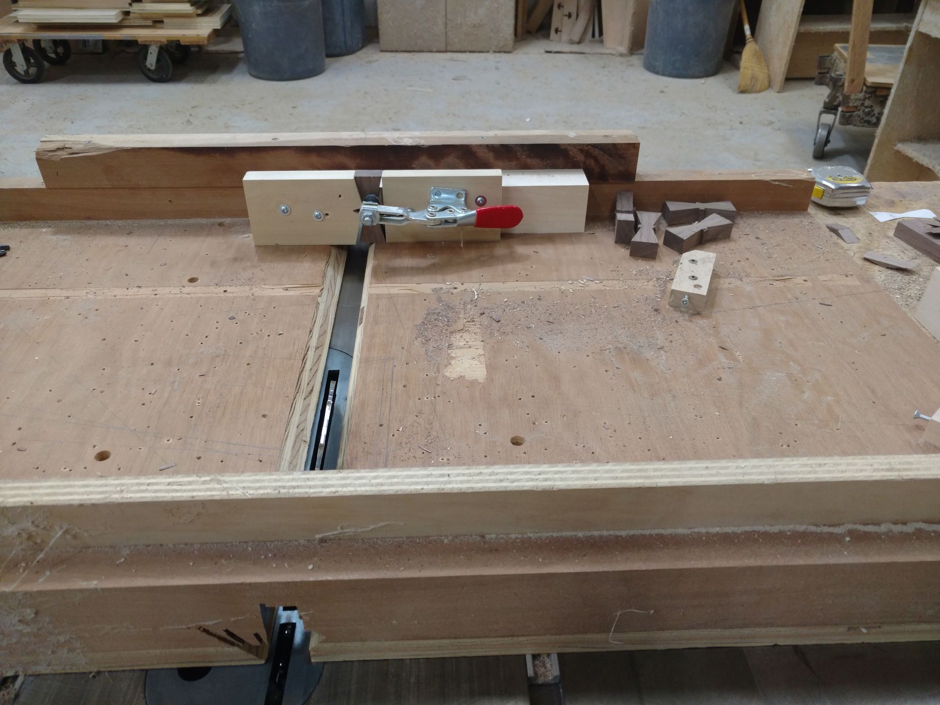 A wooden workshop table with a jig holding a wooden piece over a saw blade, ready for cutting. The jig has a red lever clasp.