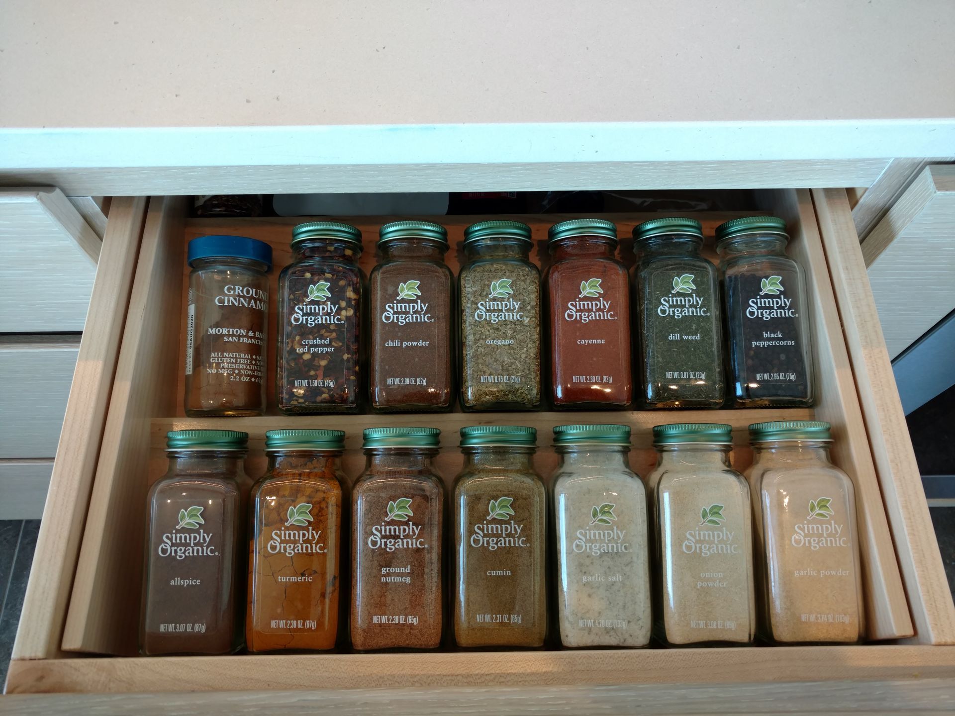 A kitchen drawer filled with spice jars of various colors, arranged on a wooden rack. The jars are labeled with green and white labels.