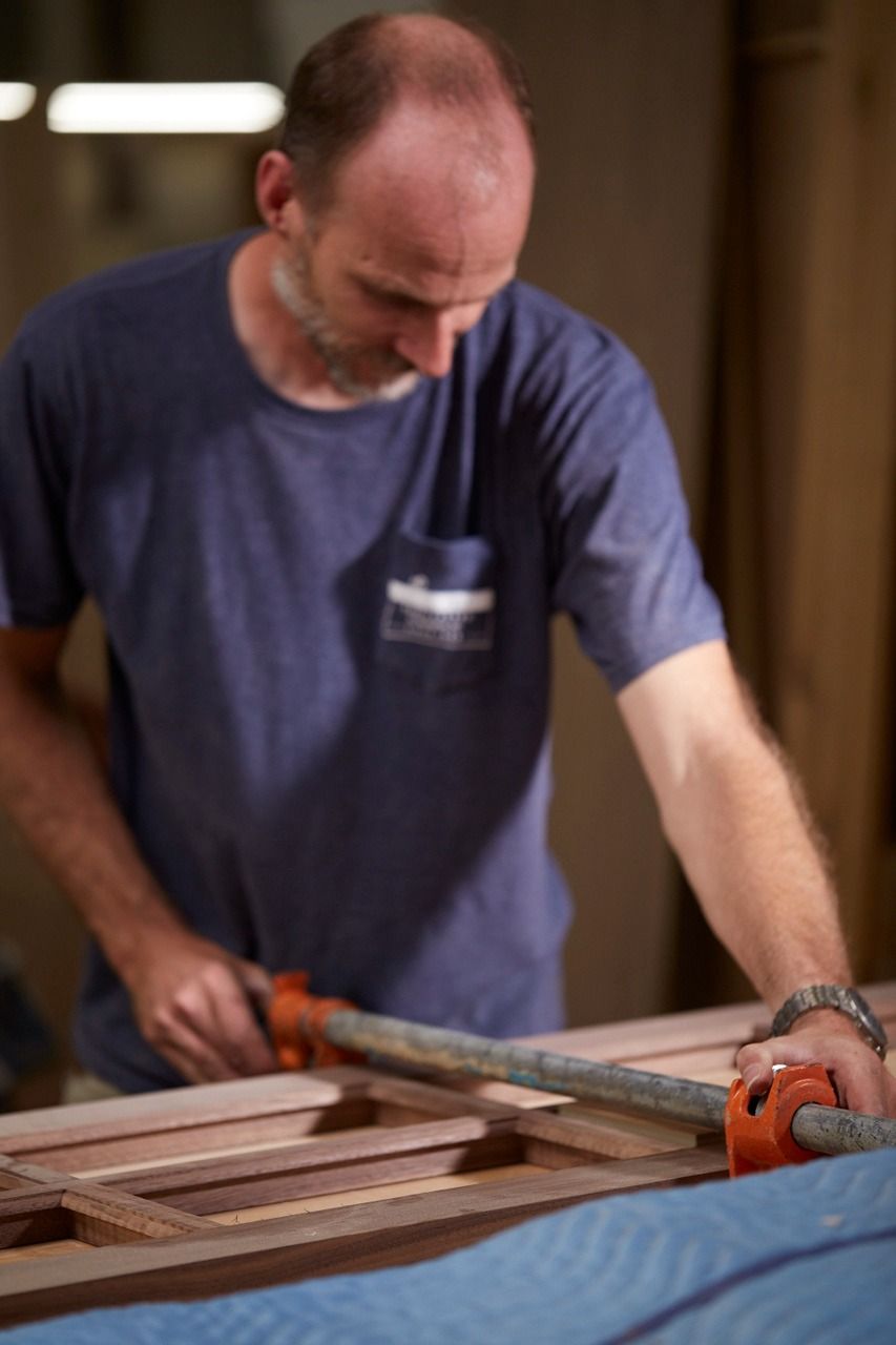 Man in blue shirt using clamps on wooden frame. He is focused, working in a workshop.