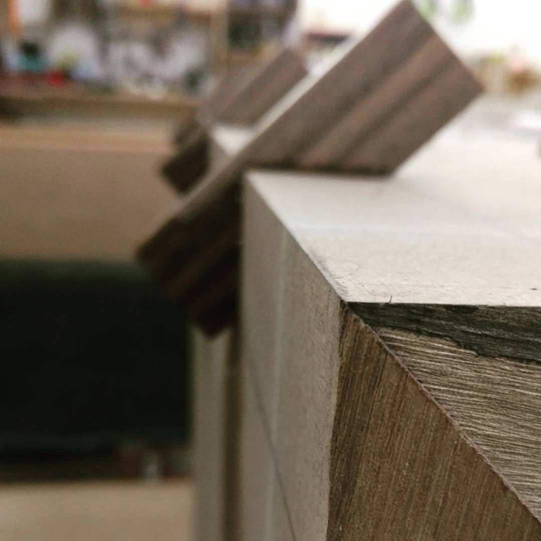 Close-up of a wooden furniture corner with angled edges, showcasing precise joinery. In the background, more wood pieces are slightly blurred.