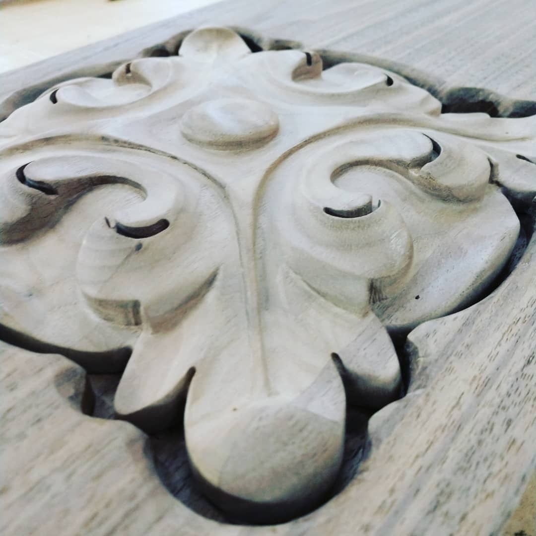 Close-up of a carved wooden ornament. A central medallion shape is surrounded by swirling, stylized leaf patterns.