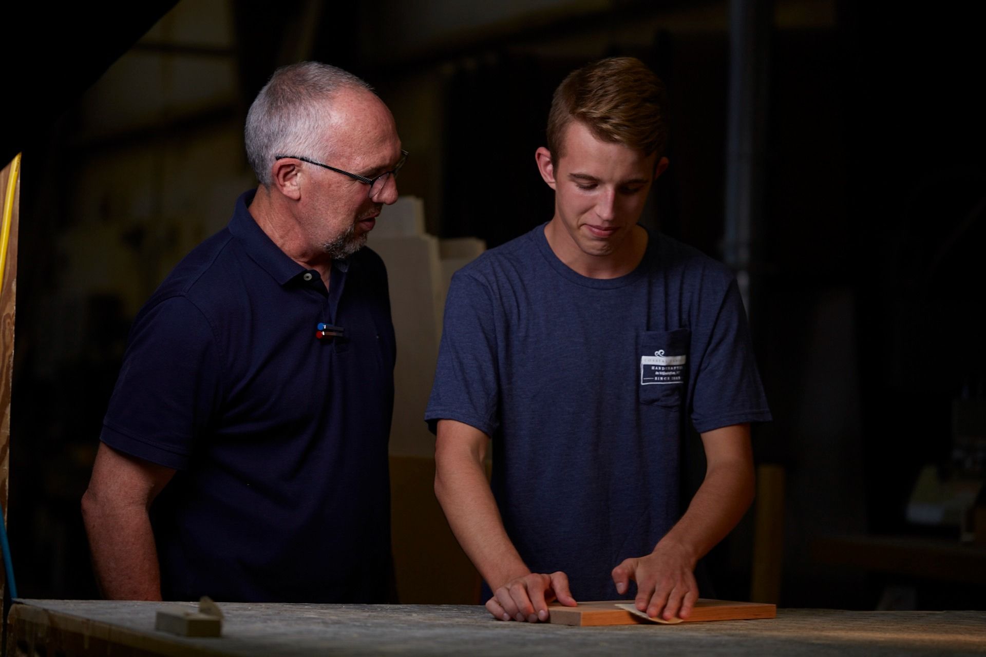 An older man watches a young man sanding wood at a workbench in a workshop. Both are wearing blue shirts.