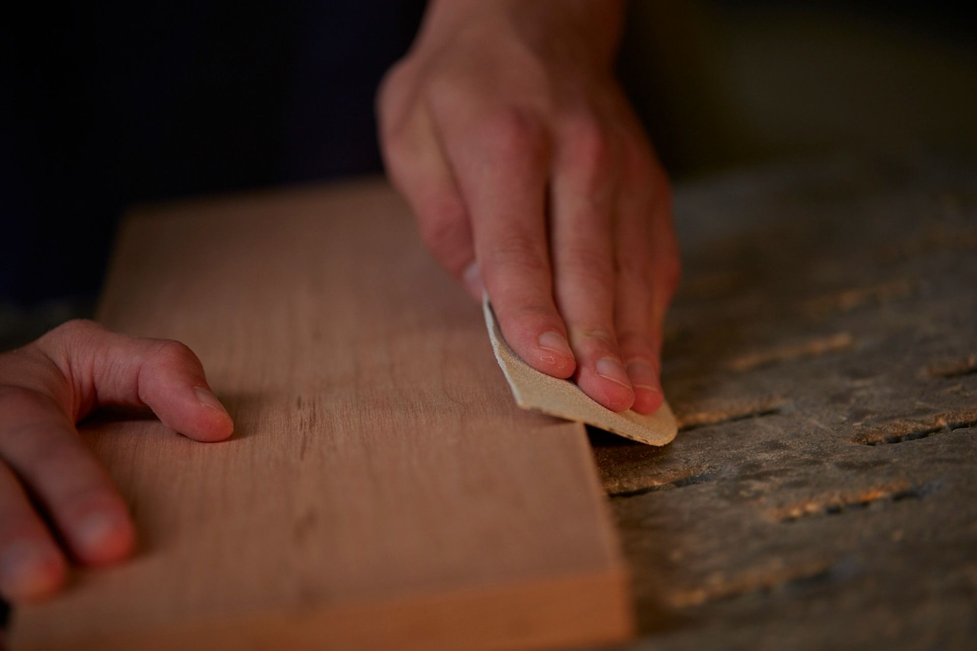 Hands sanding a wooden plank on a weathered surface. The wood is light brown; the background is dark.
