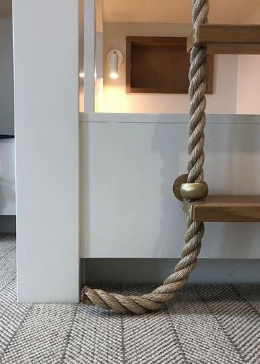 Rope ladder with wooden steps descends indoors. The rope rests on a gray and white patterned rug.