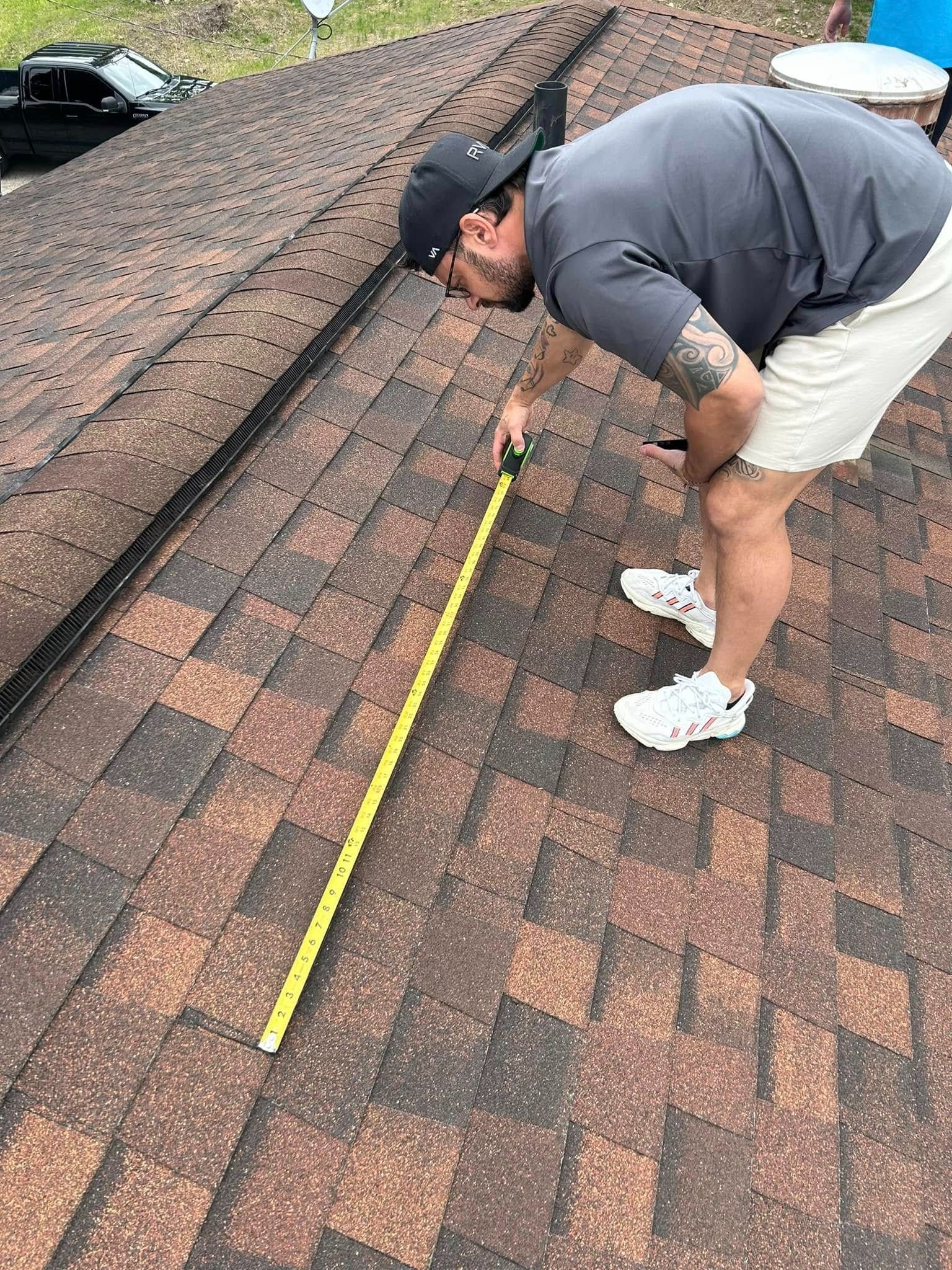 Man on a roof measures with a tape measure. Dark brown and tan shingles, gray shirt, white shorts, sneakers.