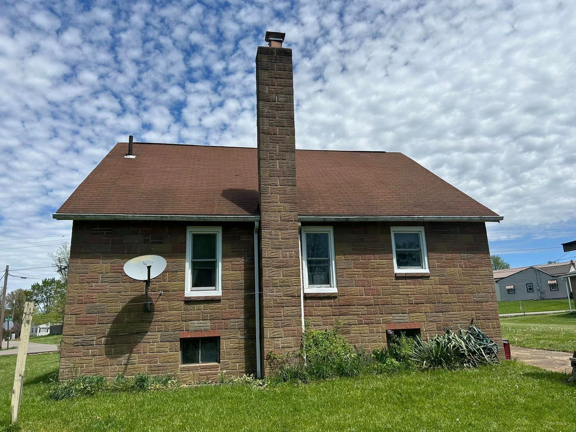 Brick house with brown roof, tall chimney, and three windows against a cloudy sky.