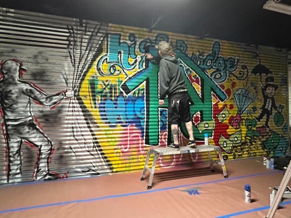 Person on stepladder paints a large, colorful mural on a corrugated metal wall. A figure pulls back a curtain on the left.