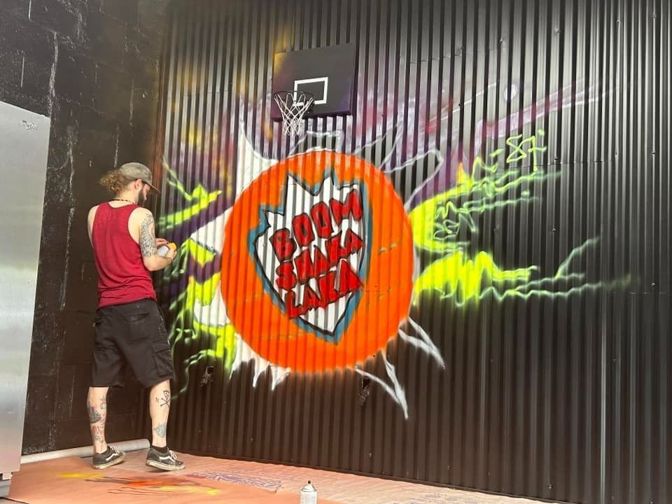 Person spraying graffiti on a wall, basketball hoop above a shield design.