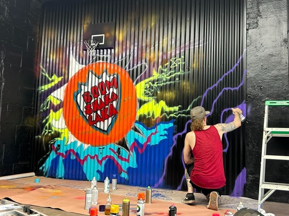 Person spray-painting purple lightning on a corrugated metal wall mural featuring a basketball hoop and colorful graffiti.