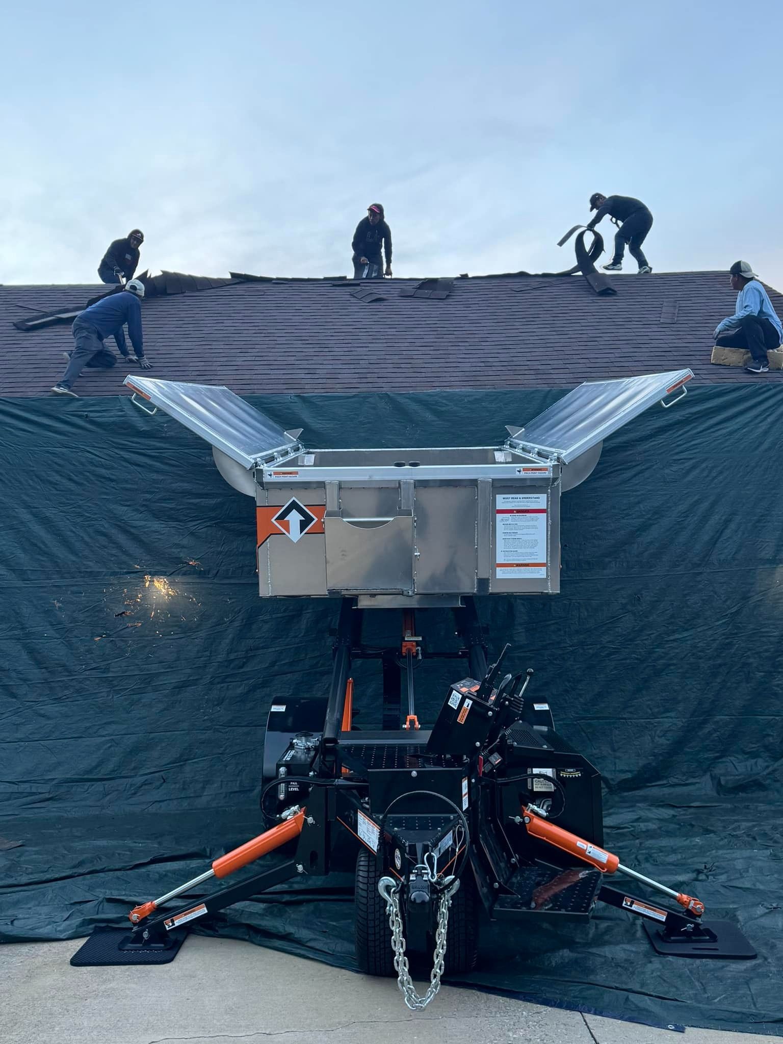 Workers on a rooftop, using a mechanical lift with a bin. Overcast sky.