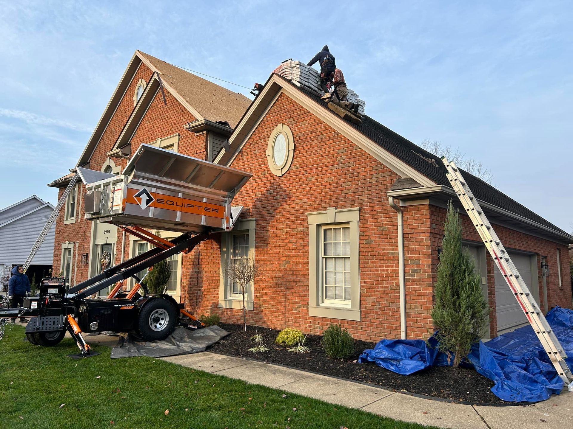 Roofers working on a brick house using a material lift. A ladder is propped against the roof.