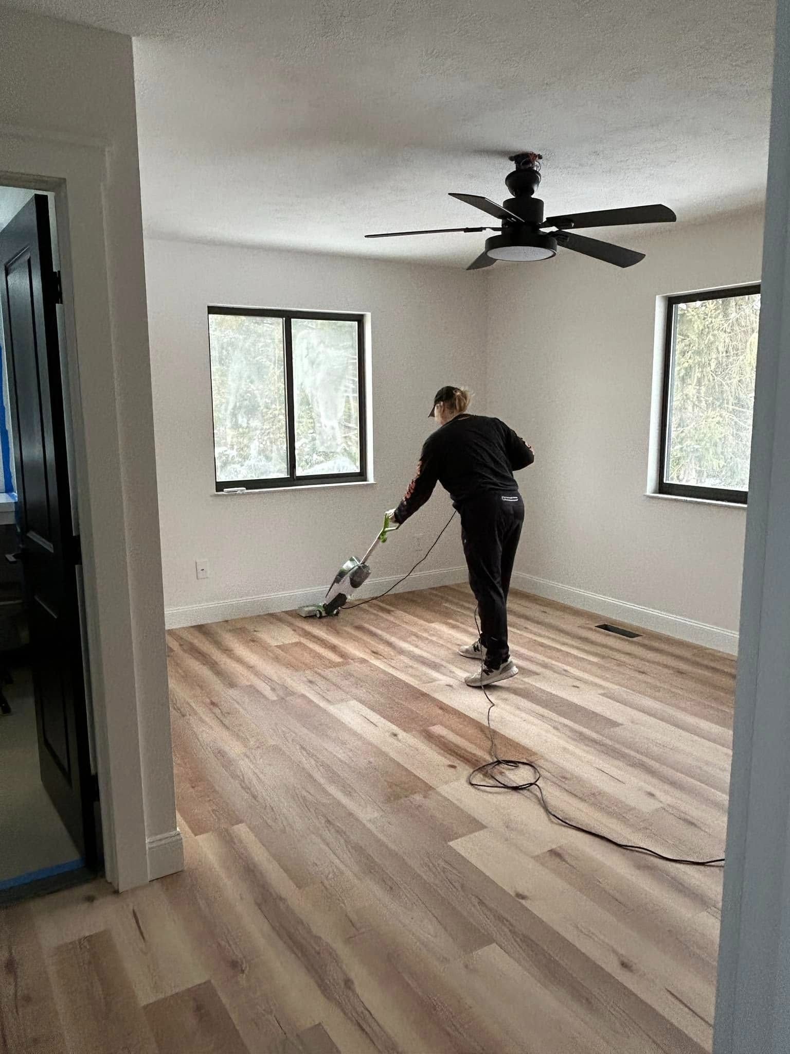 Person vacuuming wood floor in a room with black-framed windows, ceiling fan, and open door.