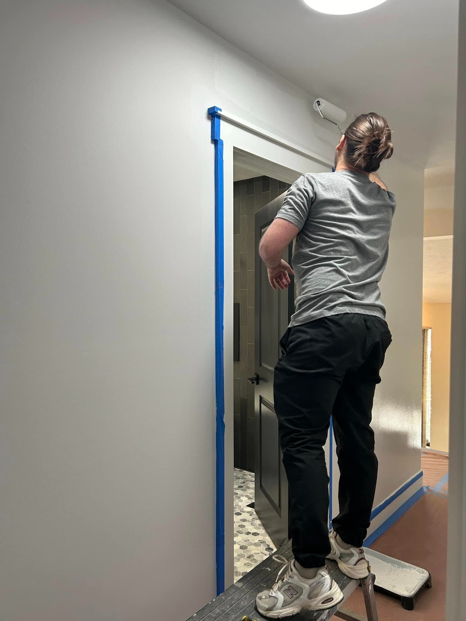 Person painting a white wall with a roller, using a step stool in a hallway, door frame taped.