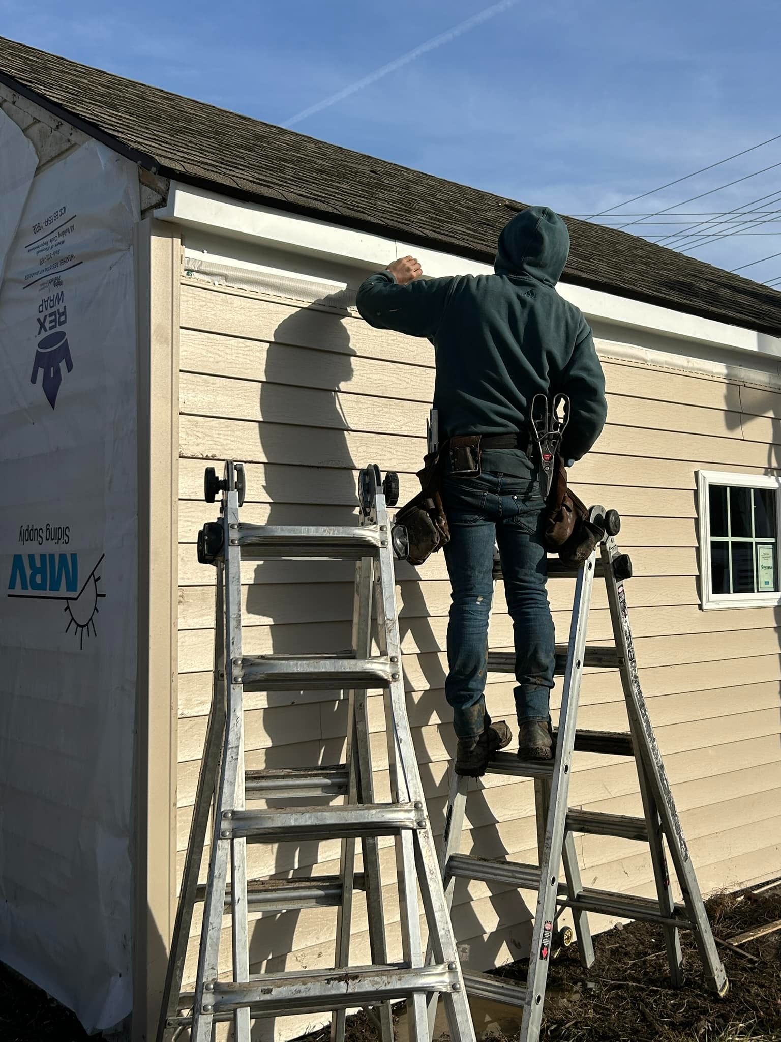Person on ladders installing siding on a building with a white window, sunny day.