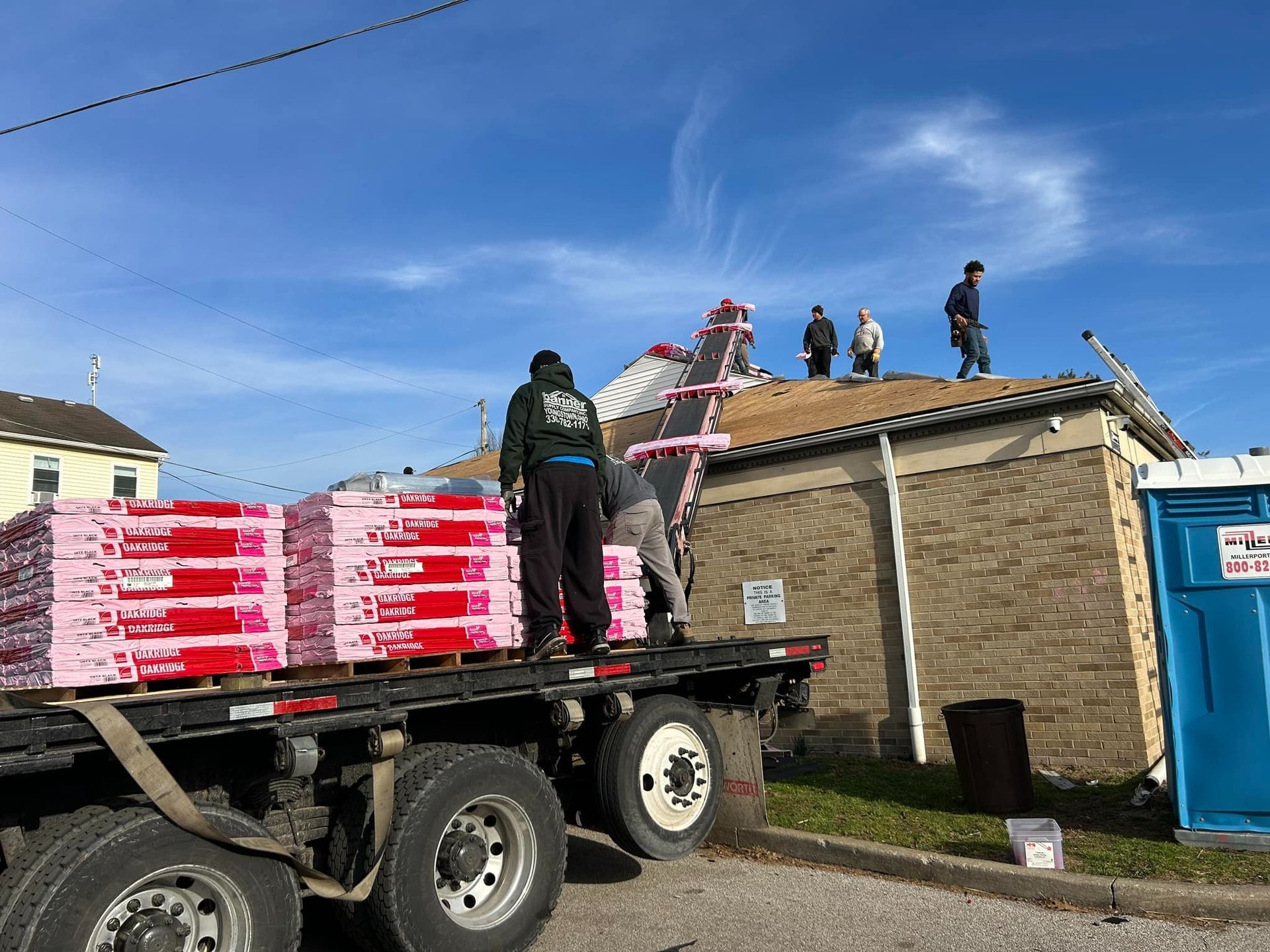 Roofing workers unloading shingles from a flatbed truck onto a building's roof under a blue sky.