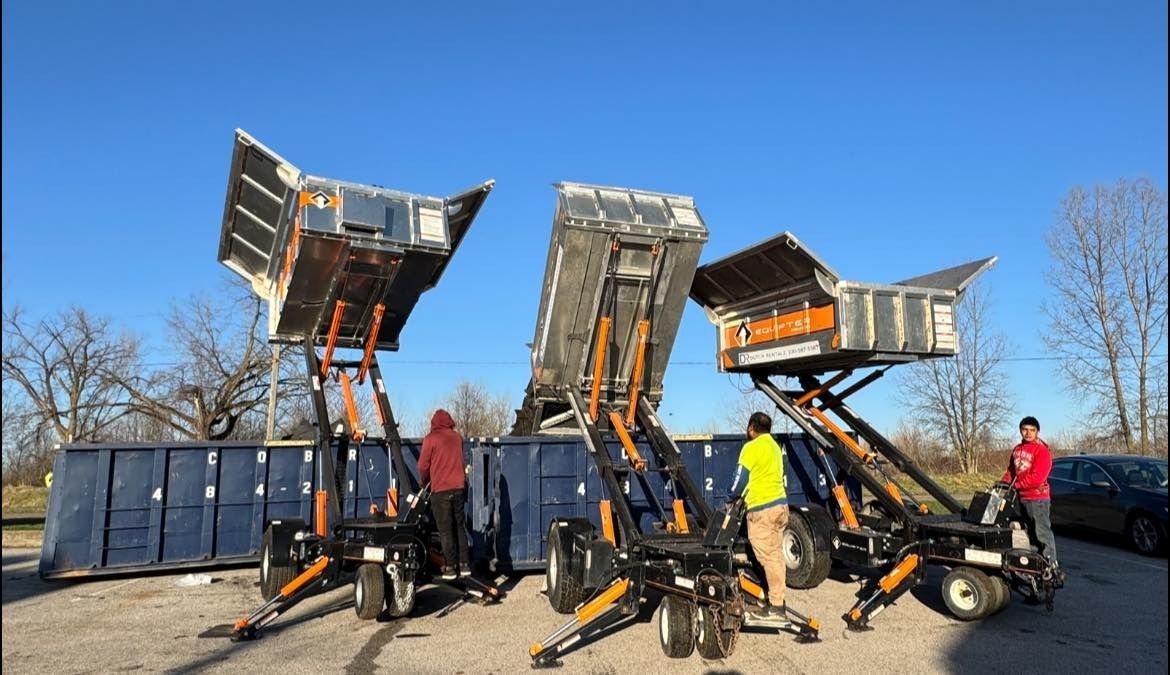 Three dumpster-loading lift machines with raised containers; three people near them on a sunny day.
