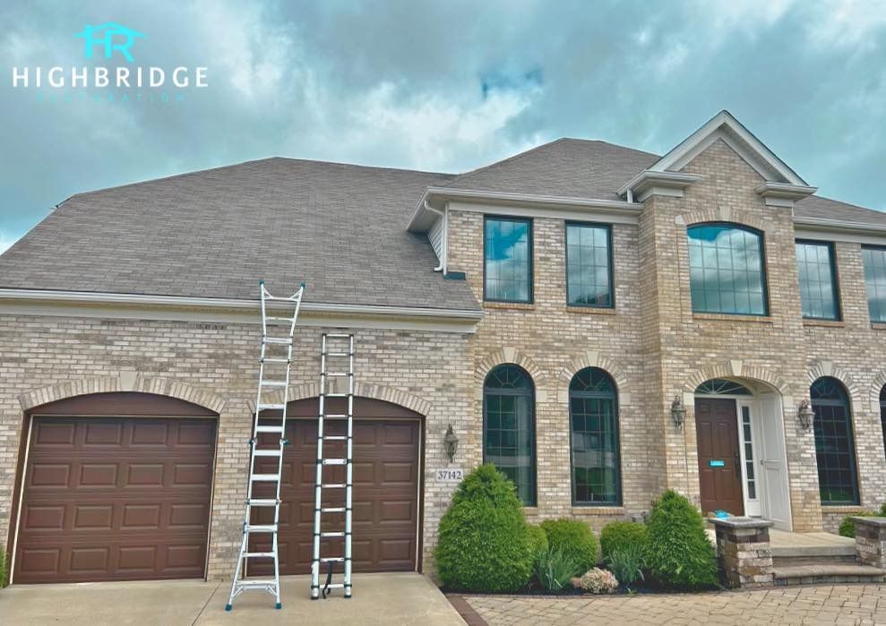 Two ladders against a house with a tan brick exterior, brown garage doors, and a brown shingle roof.