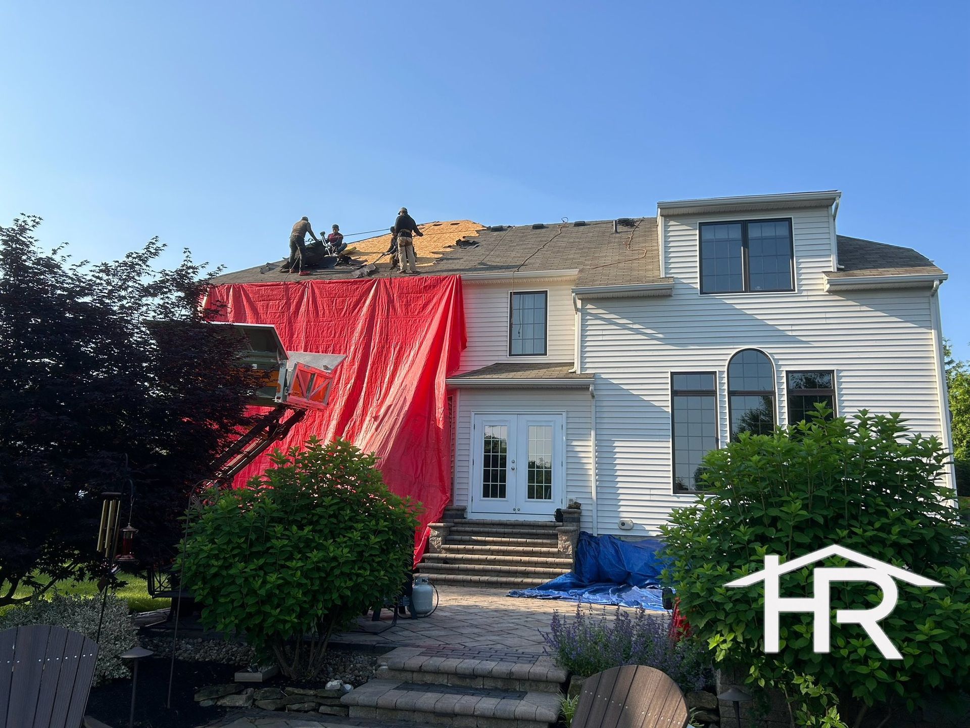 Roofers working on a house with a red tarp covering a section; blue sky in background.